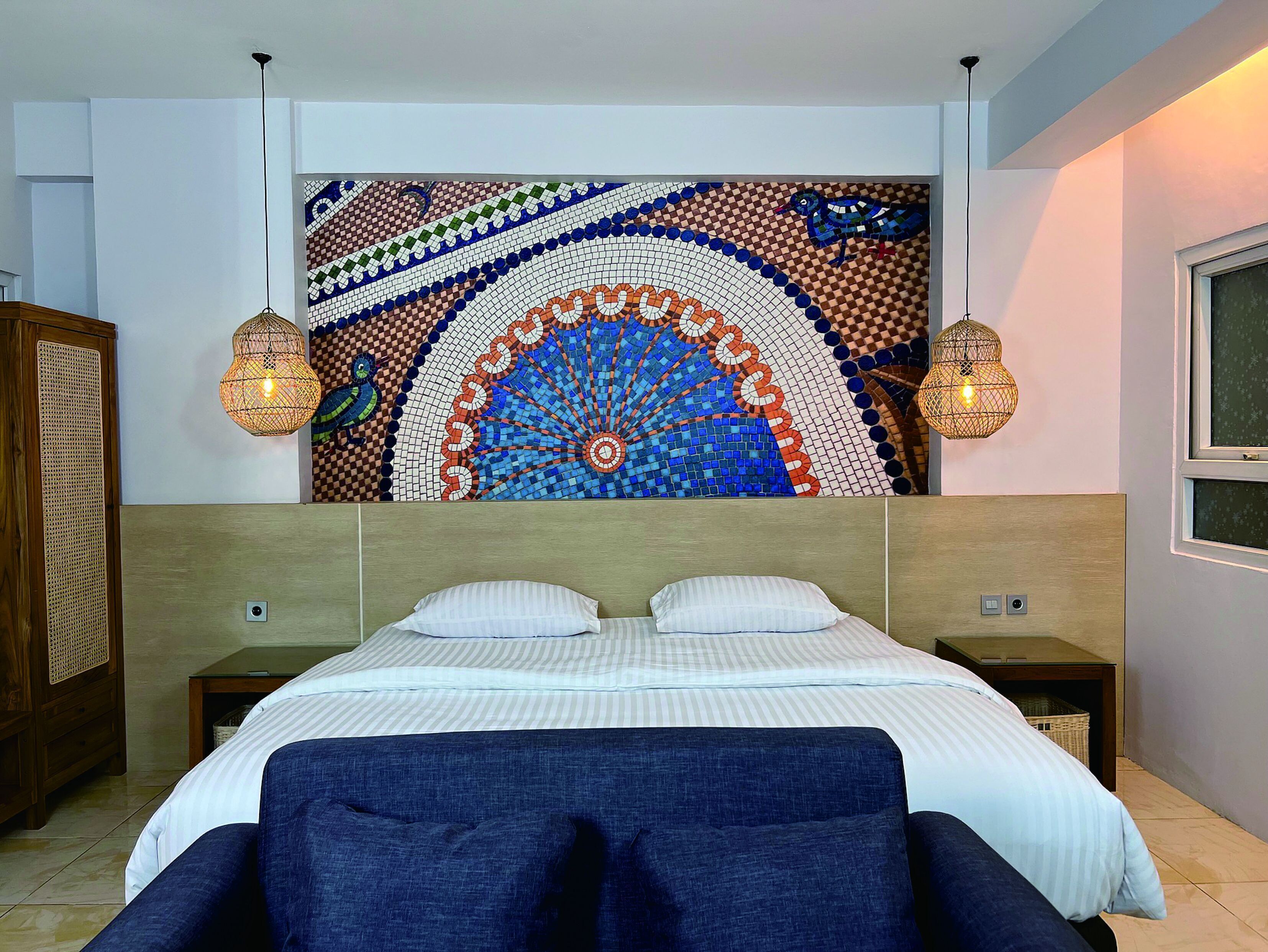 Ein Schlafzimmer mit großem, farbenfrohem Mosaikbild über dem Bett, zwei Nachttischlampen, einem Holz-Kopfteil und einem blauen, gepolsterten Sessel im Vordergrund.