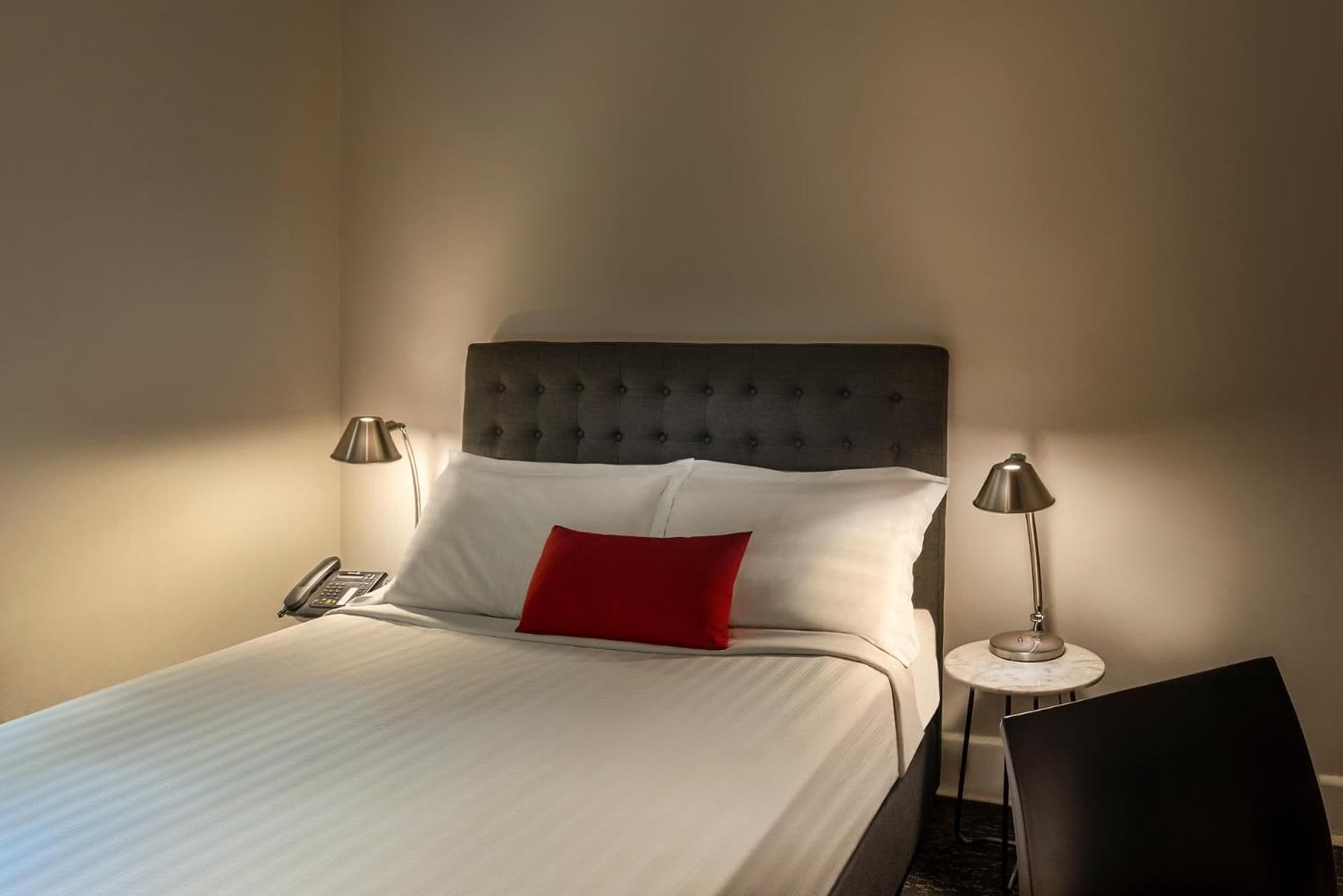 A neatly made bed with a tufted headboard, two bedside lamps, and a red pillow.