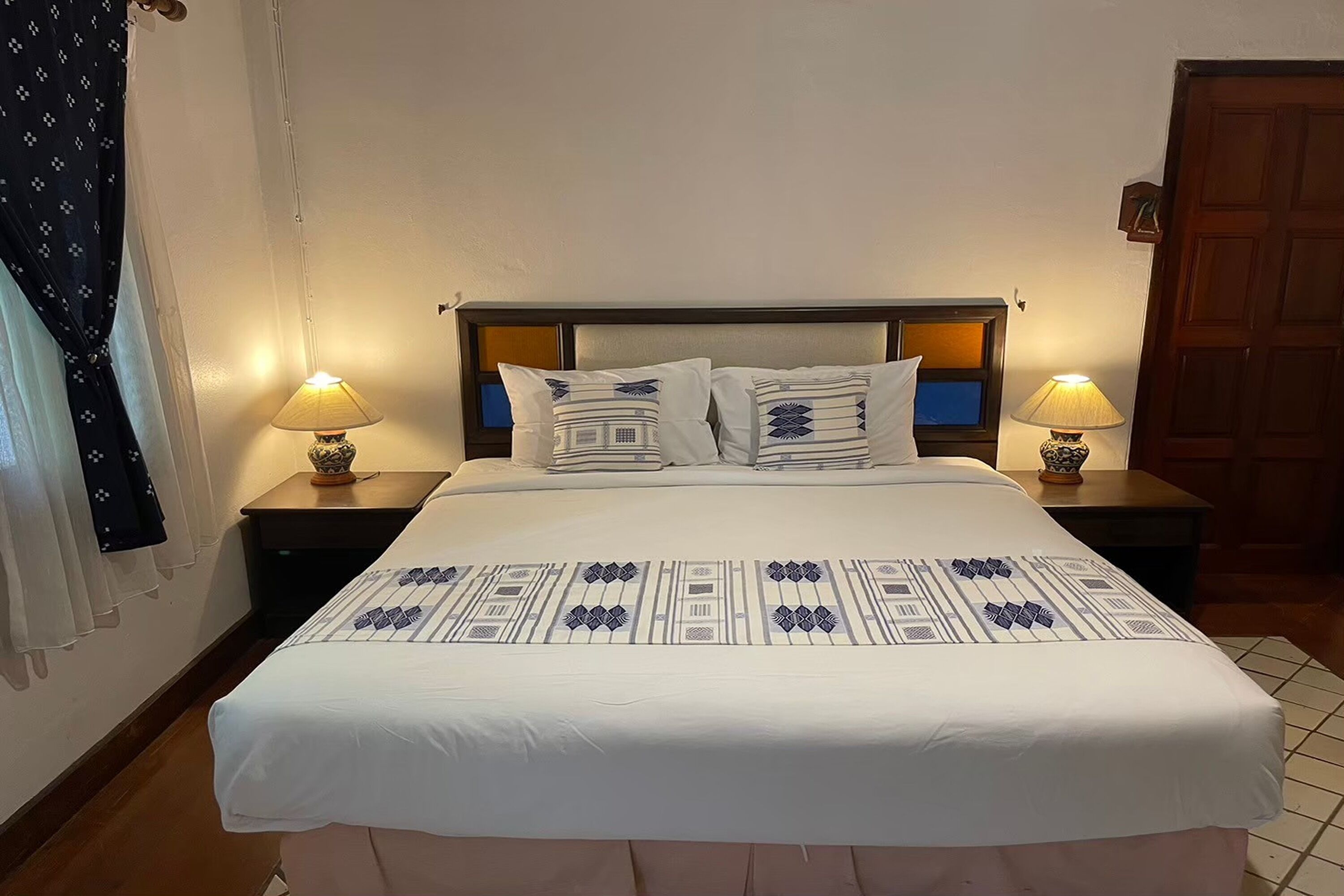 Un lit bien fait, recouvert d’une couvre-lit à motifs blancs et bleus, avec deux lampes de chevet et une tête de lit en bois.