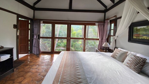 A bedroom with a large bed, a wooden floor, and a view of greenery through large windows.