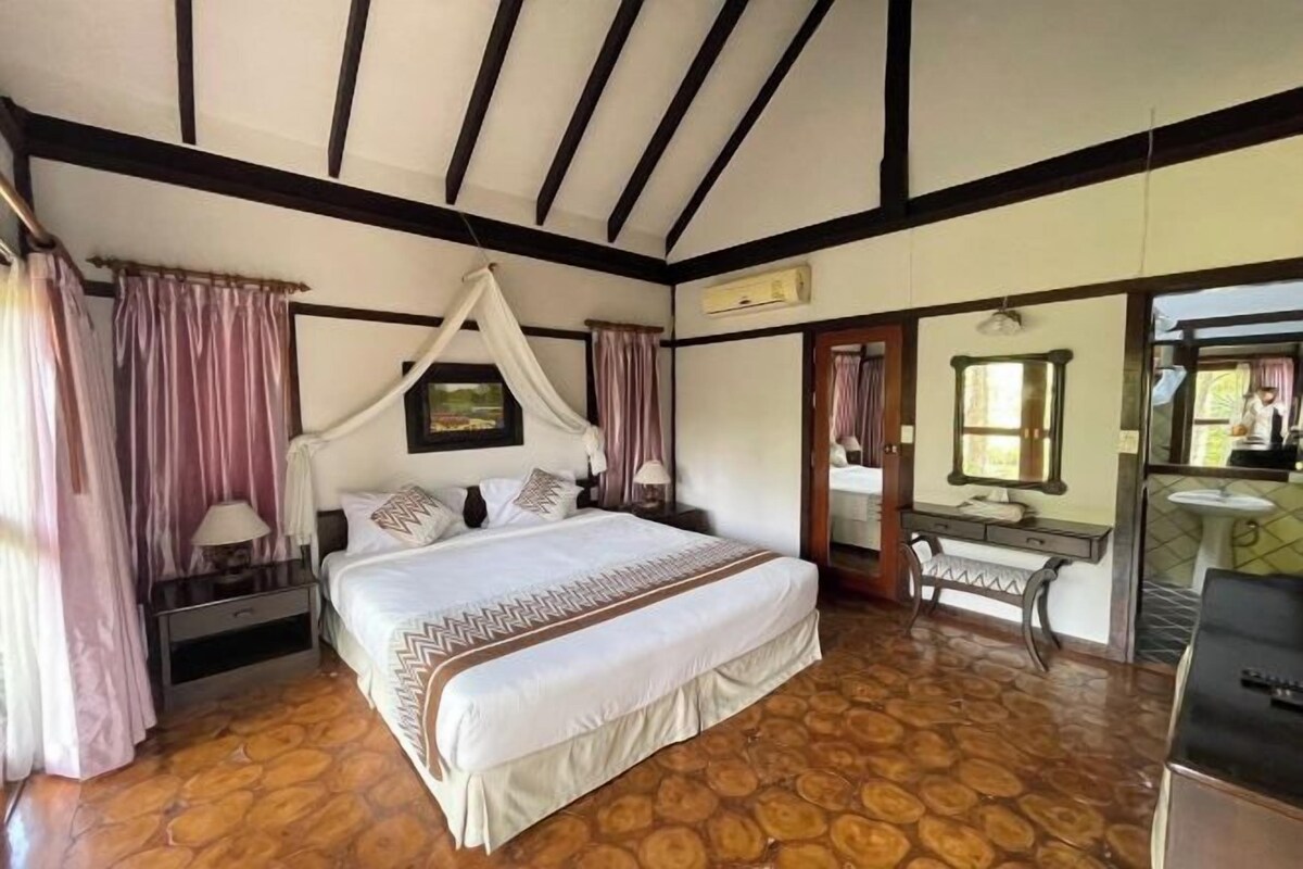 A bedroom with a large bed, a nightstand, a vanity with a mirror, and a window.
