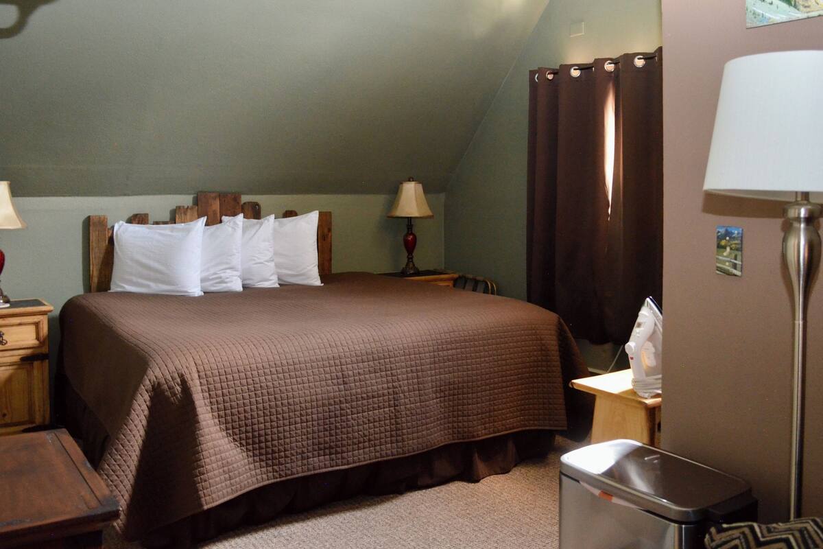 A neatly made bed with a brown quilt, two bedside lamps, a wooden nightstand, a small trash bin, and a wall-mounted lamp.