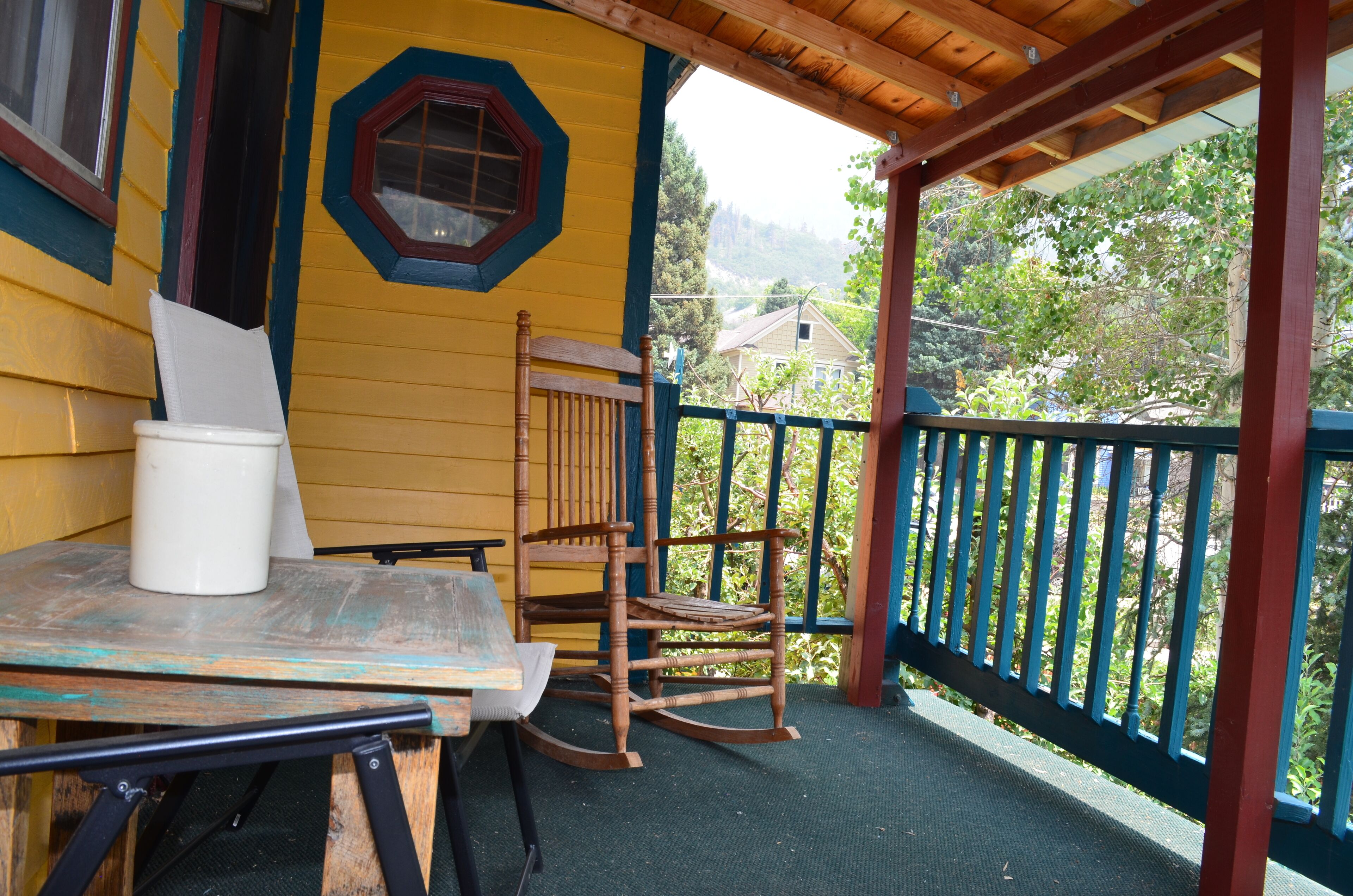 Una terraza de madera con una mesa, sillas y una mecedora, además de un ventanal con vistas a zonas verdes y edificios.