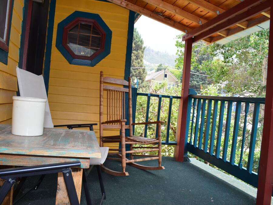 Una terraza de madera con una mesa, sillas y una mecedora, además de un ventanal con vistas a zonas verdes y edificios.