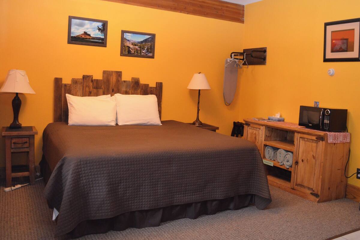 A bedroom with a large bed, two bedside lamps, a wooden headboard, a wooden cabinet with towels, and two framed pictures on the wall.