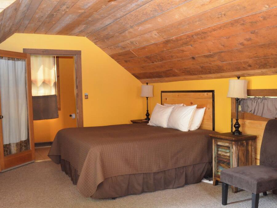 A bedroom with a wooden ceiling, a bed with a brown bedspread, two bedside tables with lamps, a chair, and a door with curtains.