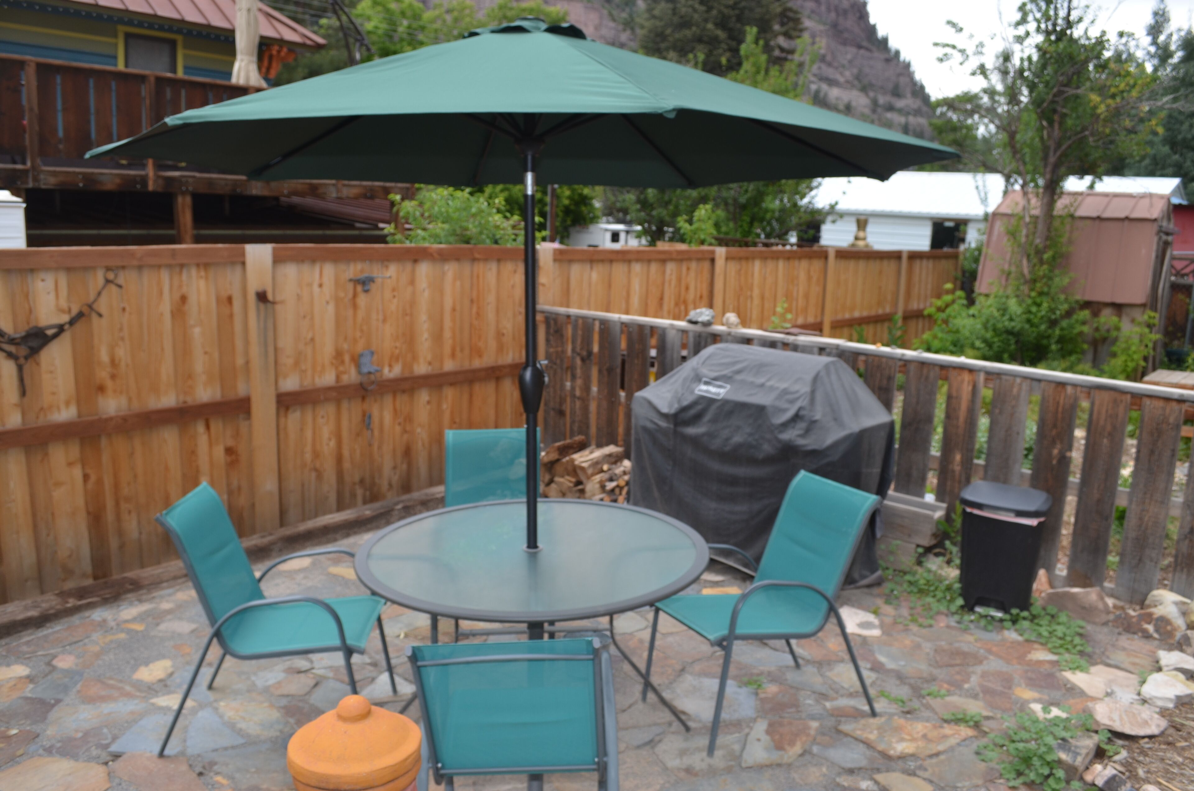 Eine Terrasse mit einem runden Tisch, vier Stühlen, einem grünen Sonnenschirm und einem mit einer Plane abgedeckten Grill.