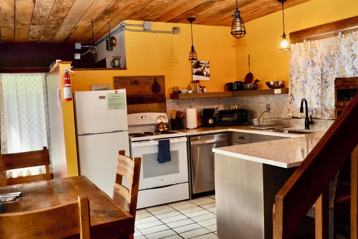 A compact kitchen with a refrigerator, stove, microwave, and sink. There is a wooden dining table and chairs, and a small island with a sink.