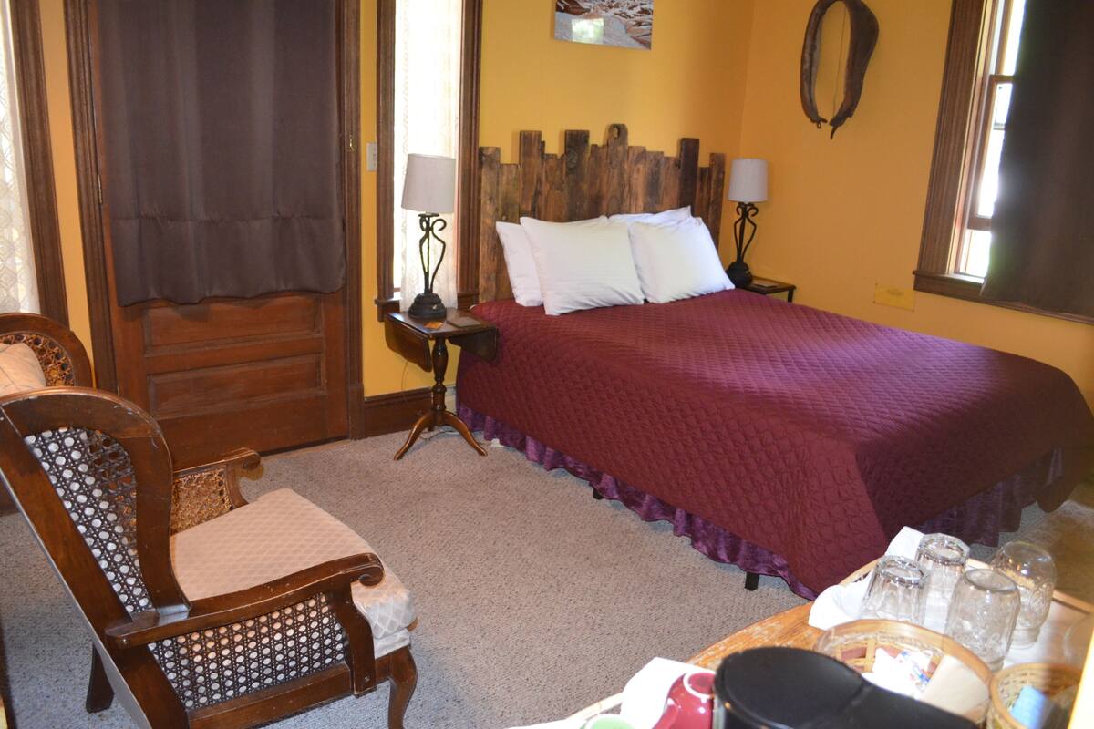 A bedroom with a wooden headboard, a bed with white and purple bedding, a nightstand with lamps, and a chair with a patterned backrest.