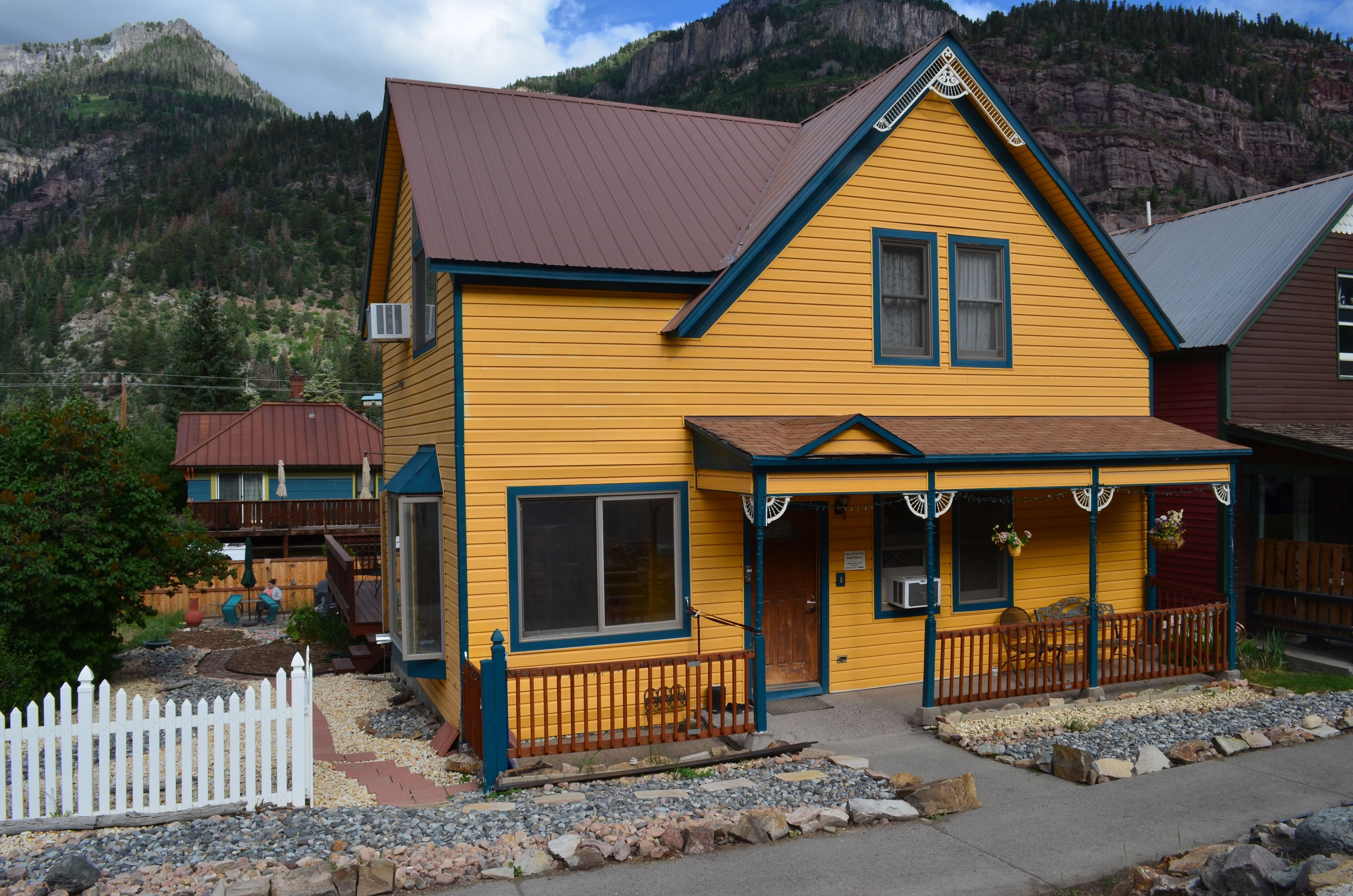 A yellow building with a brown roof, a porch with a railing, and a white picket fence.