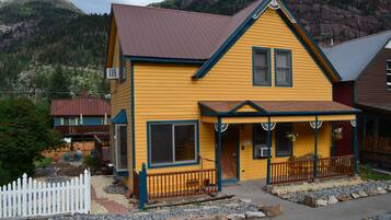 A yellow building with a brown roof, a porch with a railing, and a white picket fence.