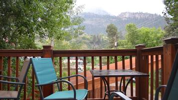 Eine Holzterrasse mit Stühlen und Tisch, mit Blick auf eine Berglandschaft, in der Ferne ist eine Straße und Fahrzeuge zu sehen.