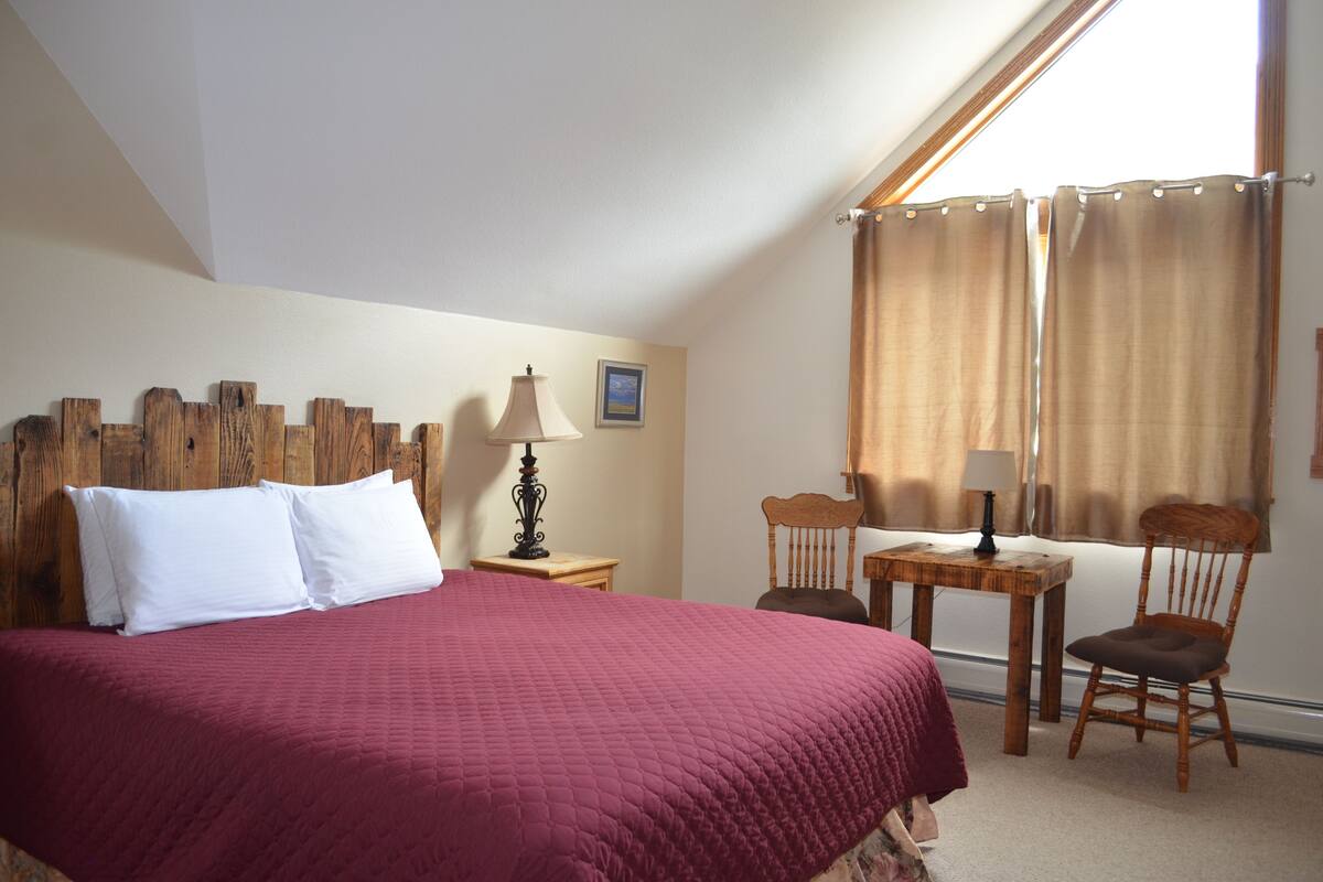 A bedroom with a large bed, a wooden headboard, a small table with two chairs, a lamp, and a window with curtains.