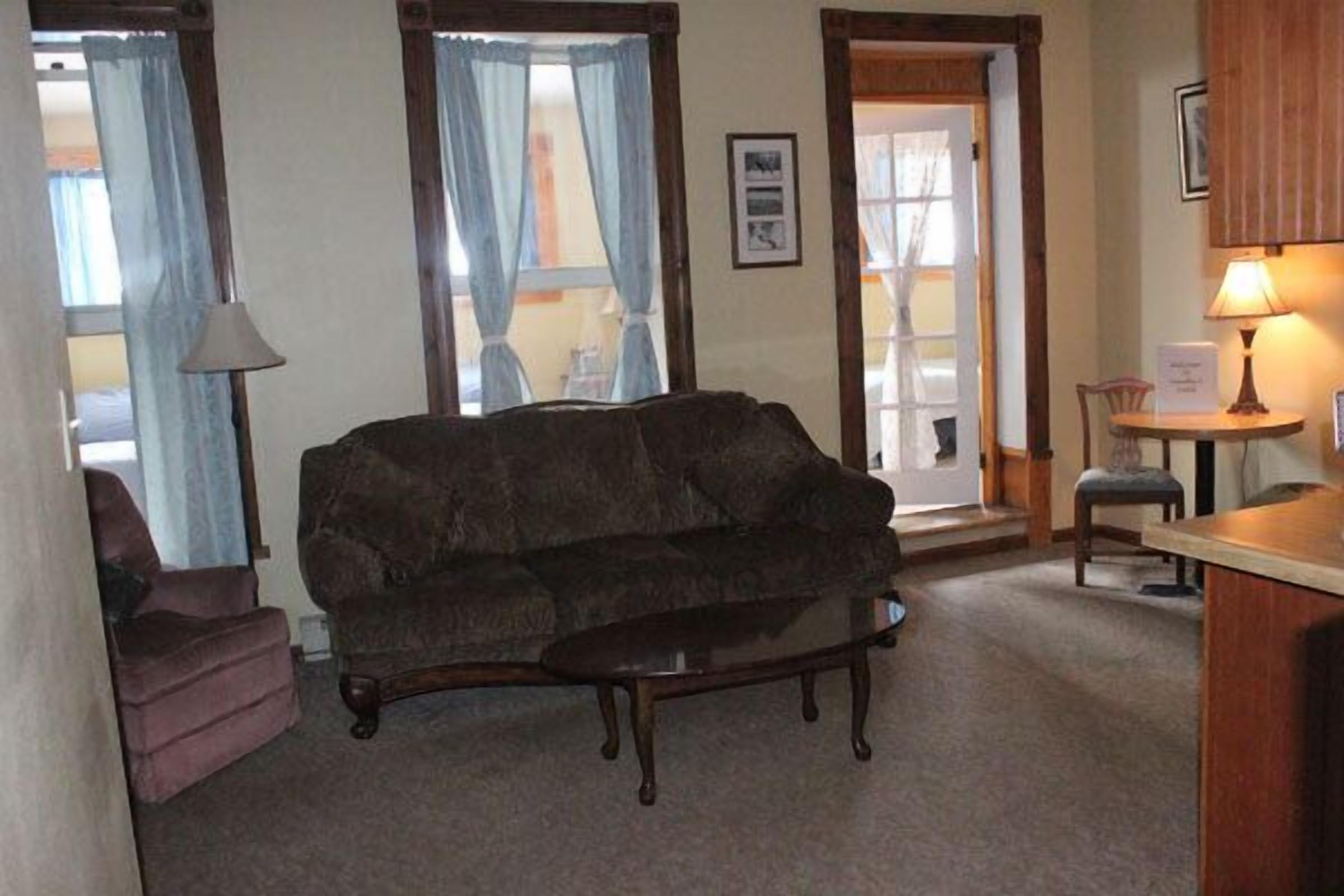 A living room with a dark brown sofa, a small armchair, a glass coffee table, and a wooden side table with a lamp.