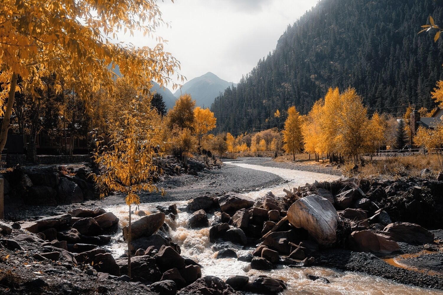 Een rivier die door een rotsachtig landschap stroomt, met herfstkleurige bomen en een berg op de achtergrond.