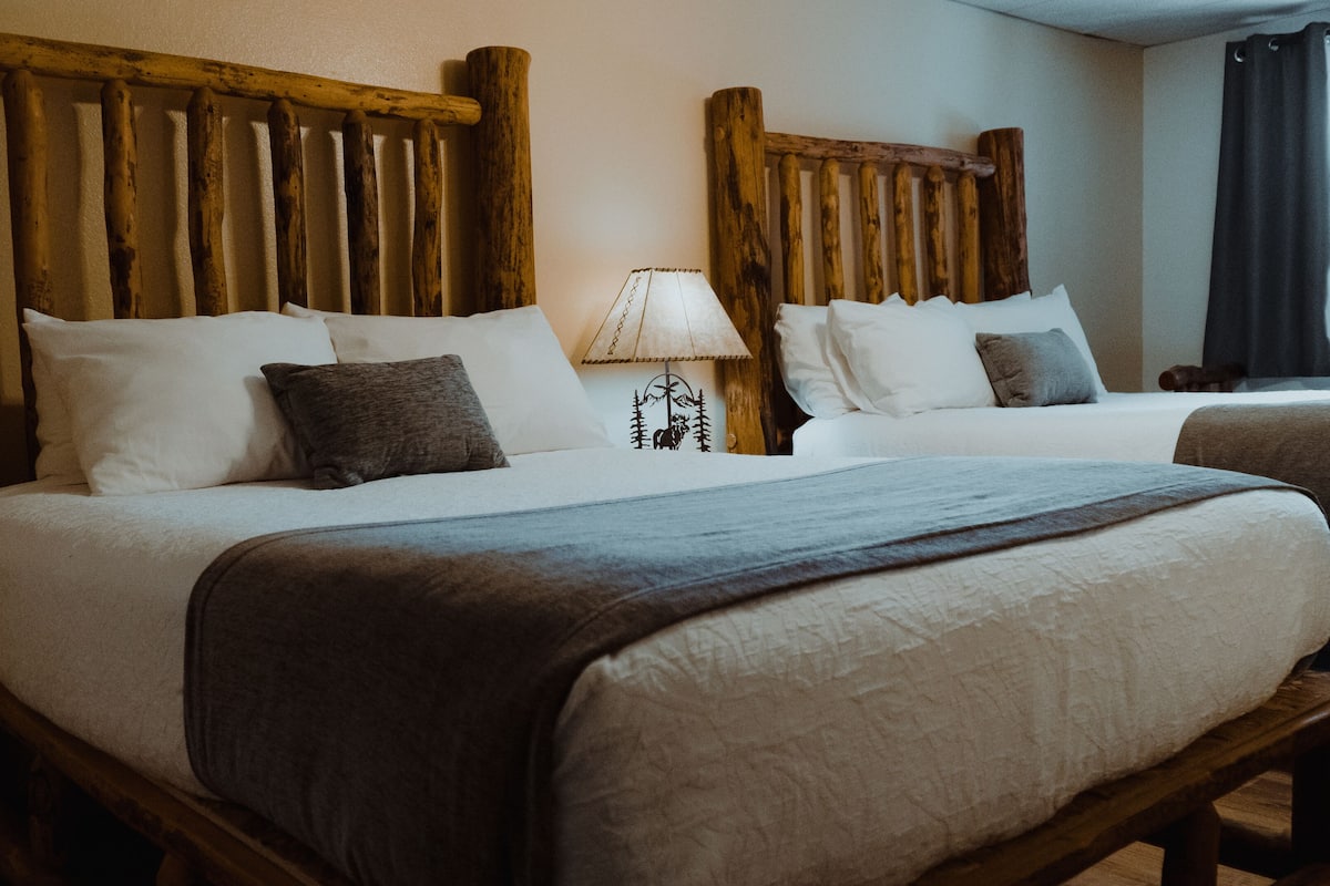 A hotel room with two beds, wooden headboards, white bedding, and a nightstand with a lamp.