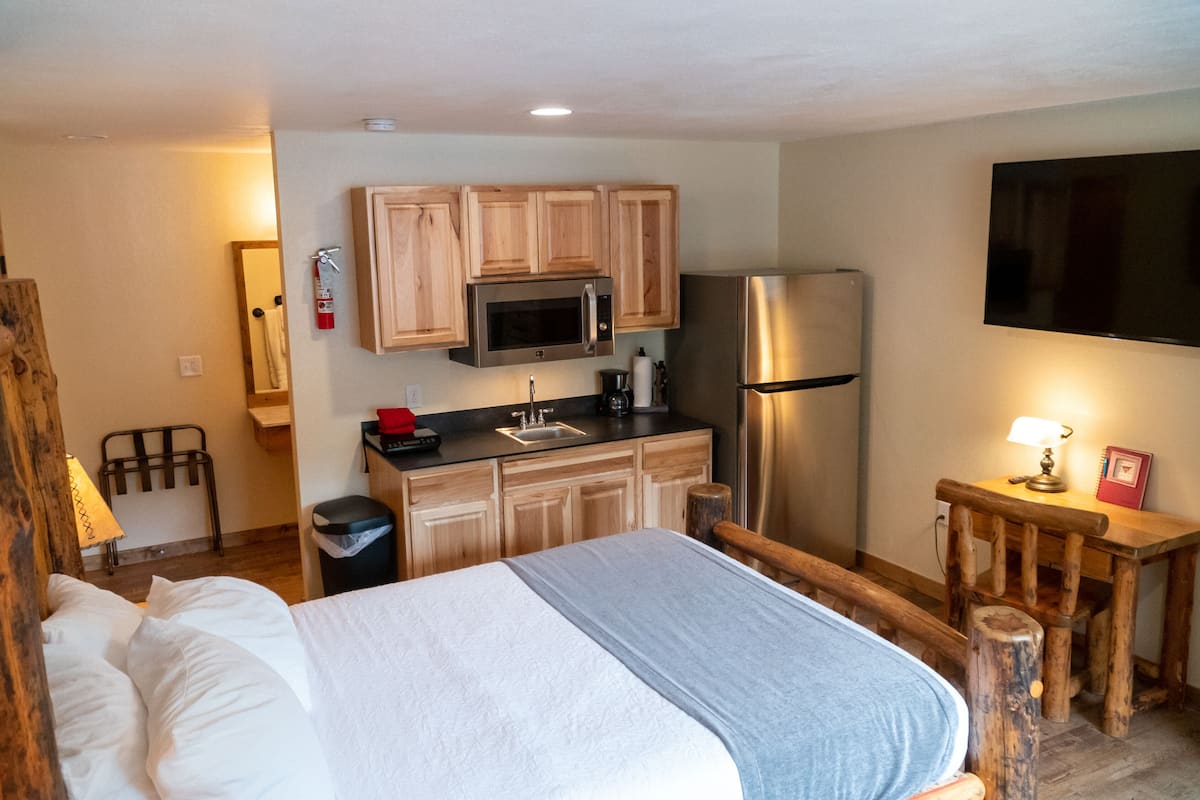 A bedroom with a bed, wooden headboard, a small desk with a lamp, and a kitchenette with a microwave, refrigerator, and wooden cabinets.