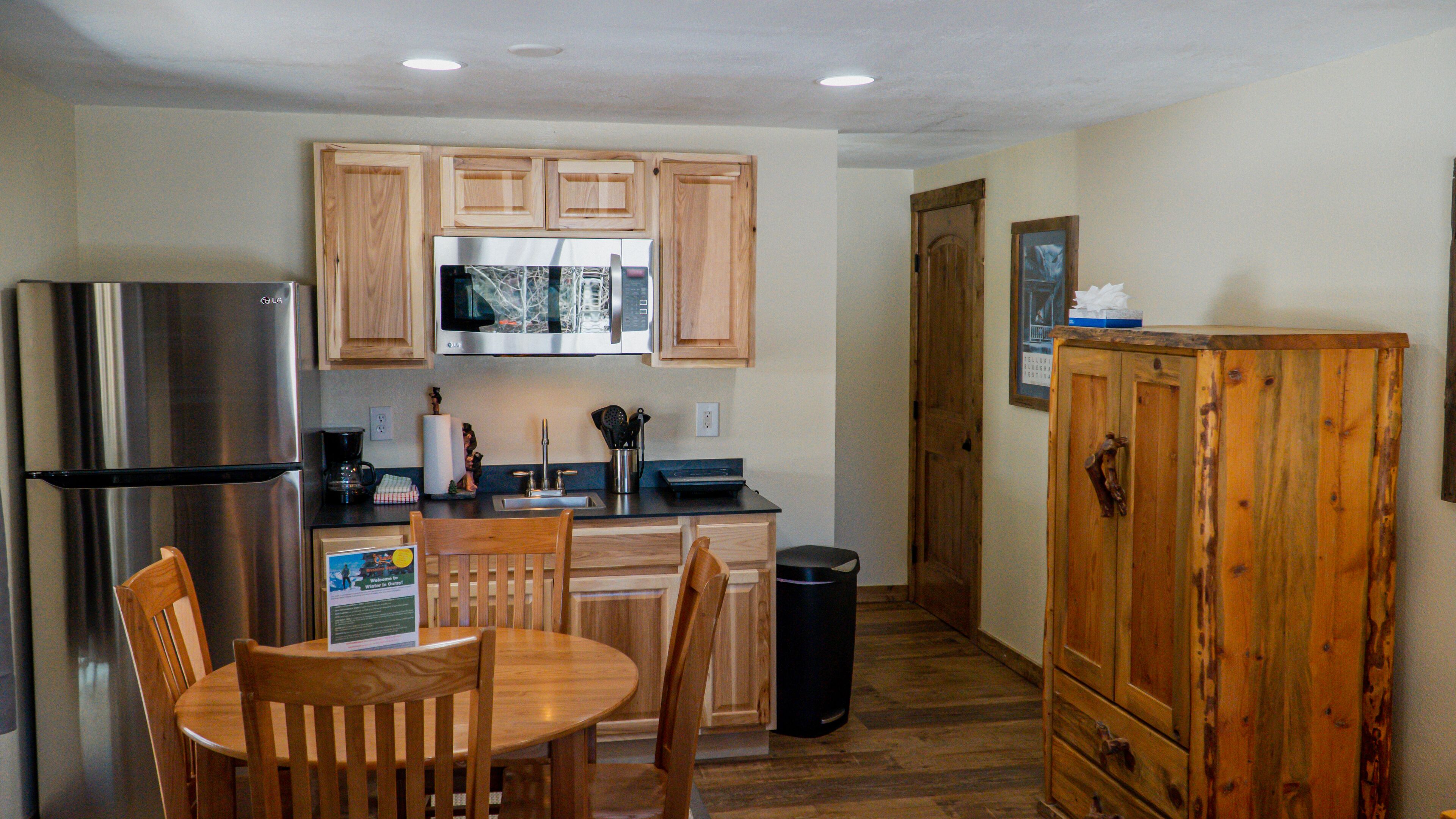 A compact kitchen with wooden cabinets, a stainless steel refrigerator, a microwave, and a sink. There is a wooden dining table with chairs, a trash can, and a cabinet with a picture on the wall.