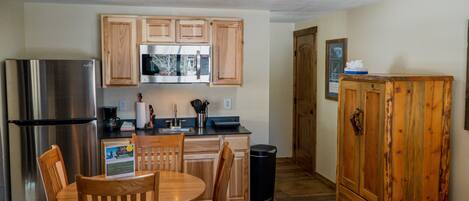 A compact kitchen with wooden cabinets, a stainless steel refrigerator, a microwave, and a sink. There is a wooden dining table with chairs, a trash can, and a cabinet with a picture on the wall.