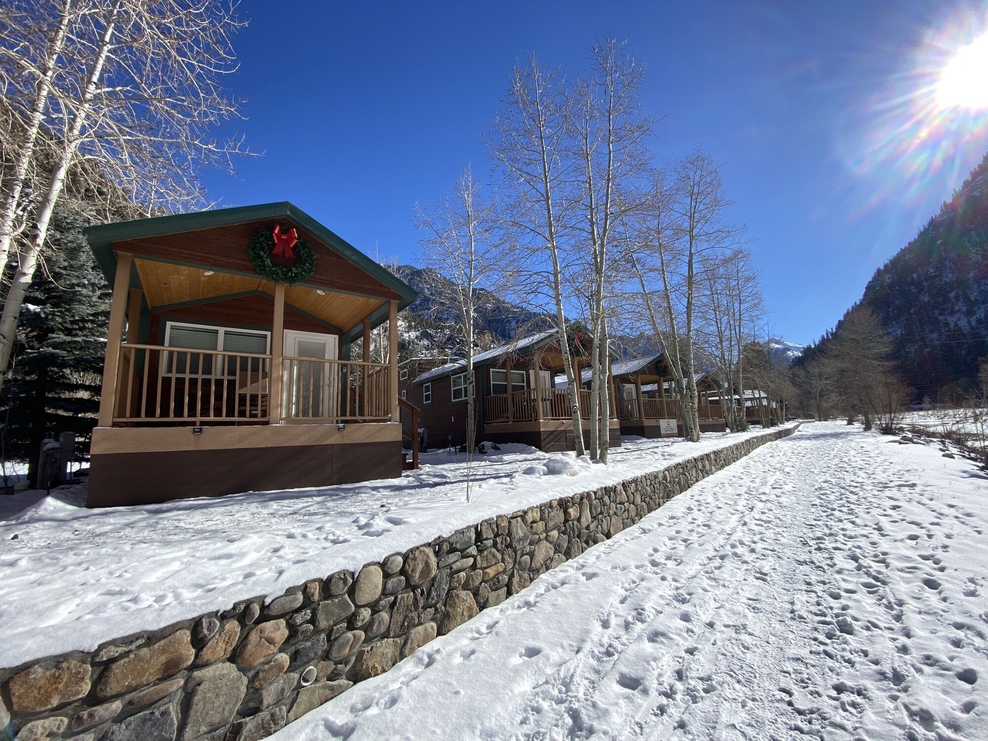 A wooden cabin with a stone retaining wall, surrounded by snow-covered ground and trees.