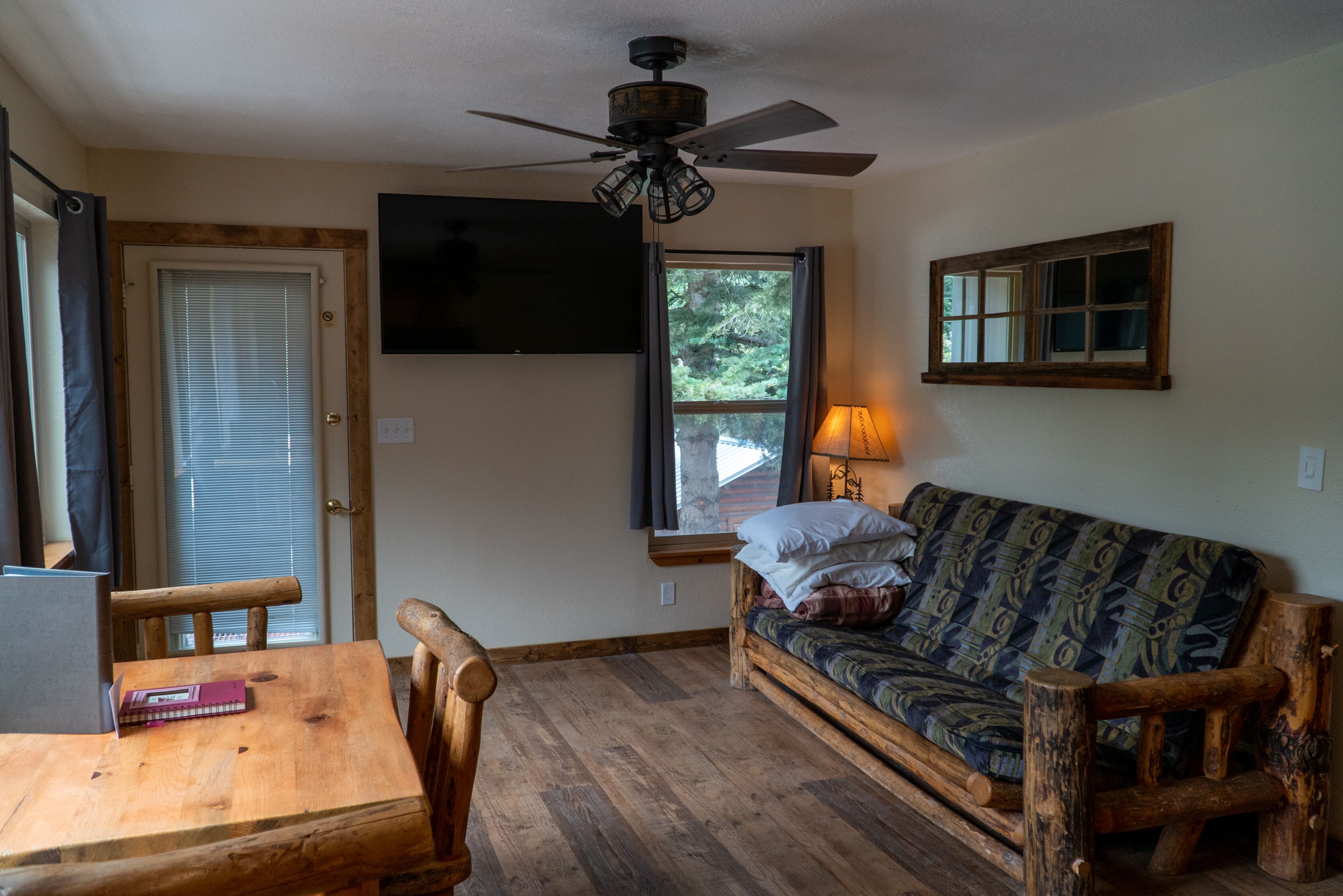 A cozy room with a wooden floor, a wooden table with a book, a wooden sofa, a ceiling fan, a TV mounted on the wall, and a window with a view of trees.