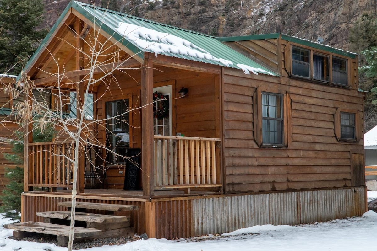 A wooden cabin with a porch, a green roof, and a view of a mountainous landscape.