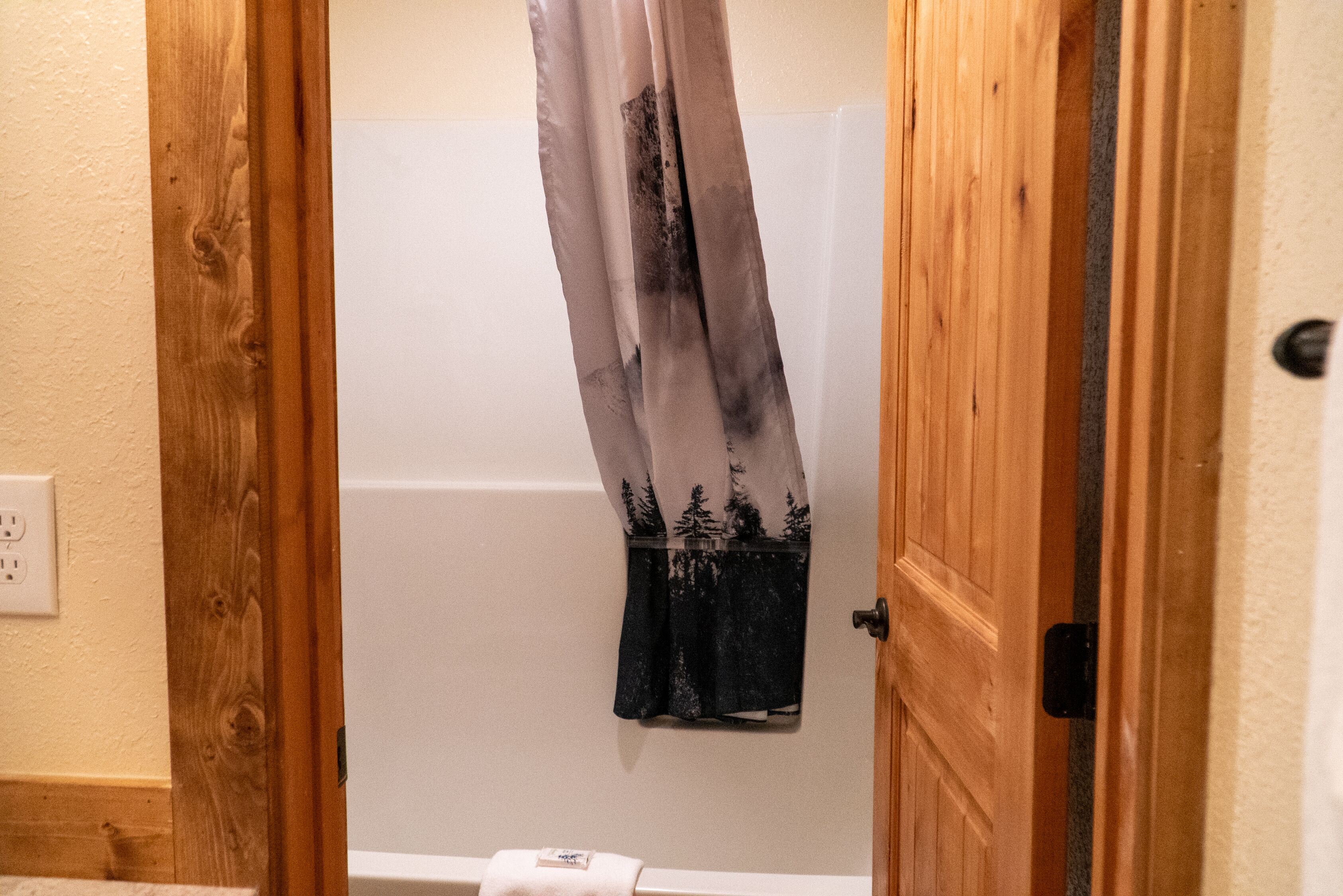 A bathroom with a wooden door, a white bathtub, and a shower curtain with a forest pattern.