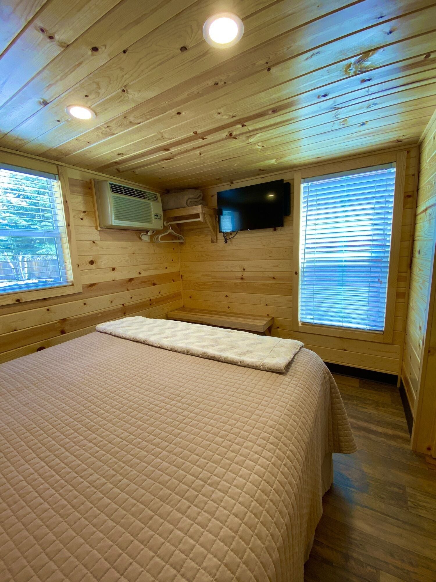 A wooden cabin room with a bed, a mounted TV, and a window with blinds.