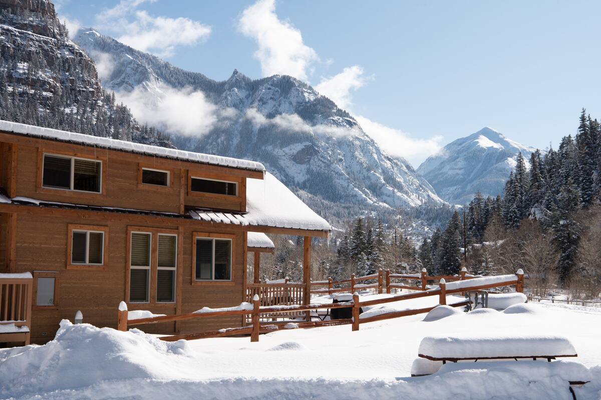 A wooden cabin with a balcony, surrounded by snow-covered mountains and trees.