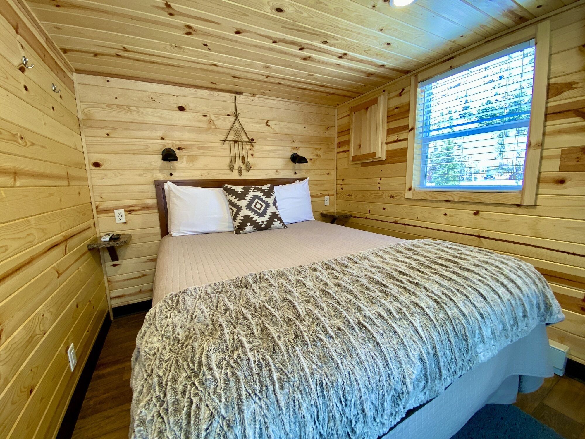 A wooden cabin bedroom with a bed, pillows, a nightstand, a window, and a wall-mounted light fixture.
