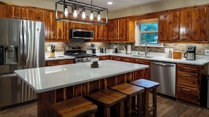A modern kitchen with wooden cabinets, a stainless steel refrigerator, a large island with stools, and a hanging light fixture. - Ouray Riverside Resort - Inn & Cabins (Ouray)