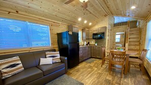 A cozy cabin interior with a wooden ceiling, a black refrigerator, a kitchen area with cabinets and a microwave, a wooden dining table with chairs, and a gray sofa with patterned cushions. - Ouray Riverside Resort - Inn & Cabins (Ouray)