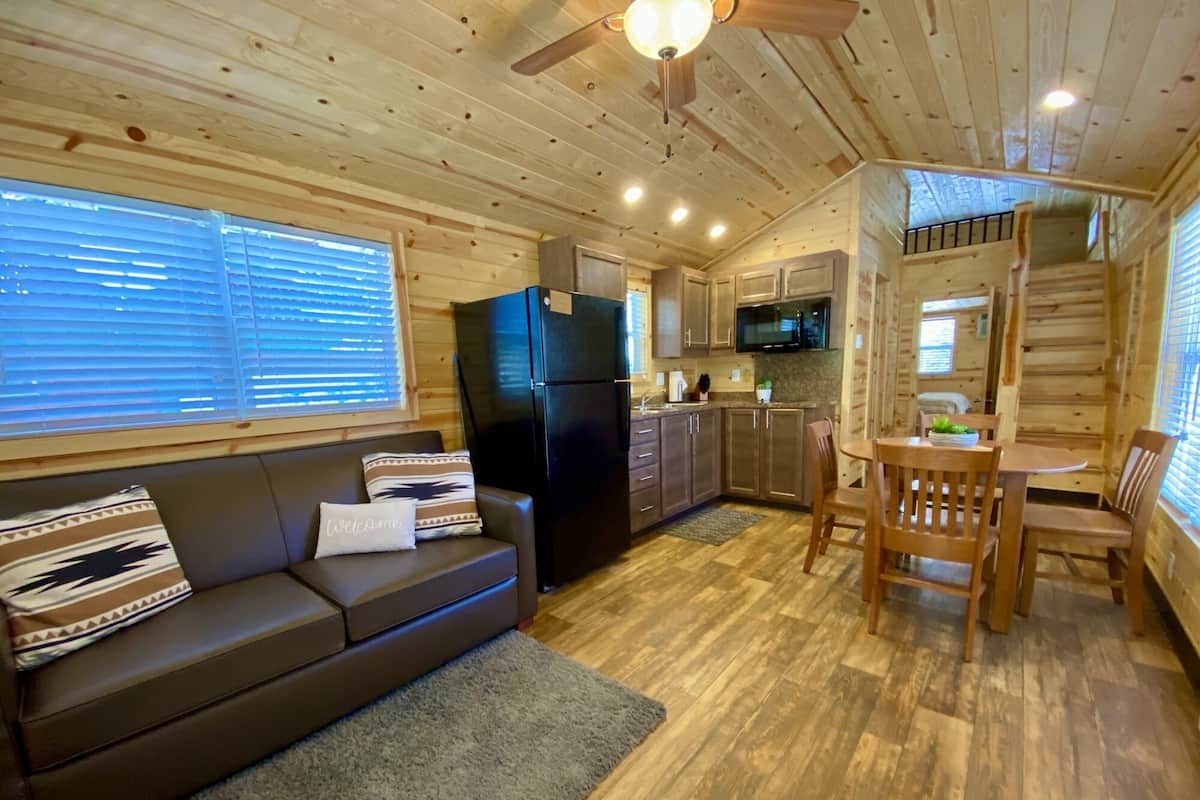 A cozy cabin interior with a wooden ceiling, a black refrigerator, a kitchen area with cabinets and a microwave, a wooden dining table with chairs, and a gray sofa with patterned cushions.