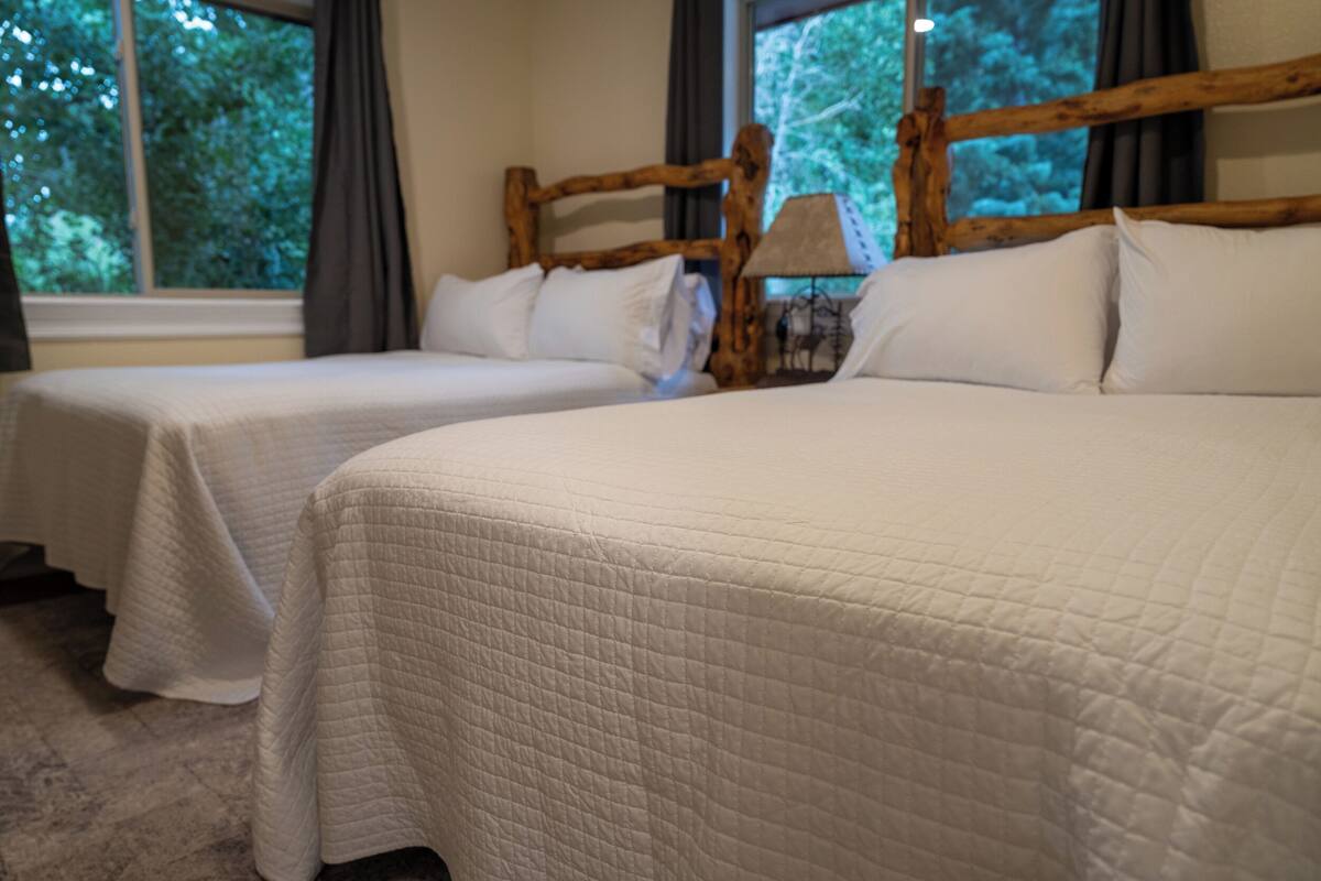 A hotel room with two beds, each with white bedding and a wooden headboard. There is a window with curtains, and a lamp on a bedside table.