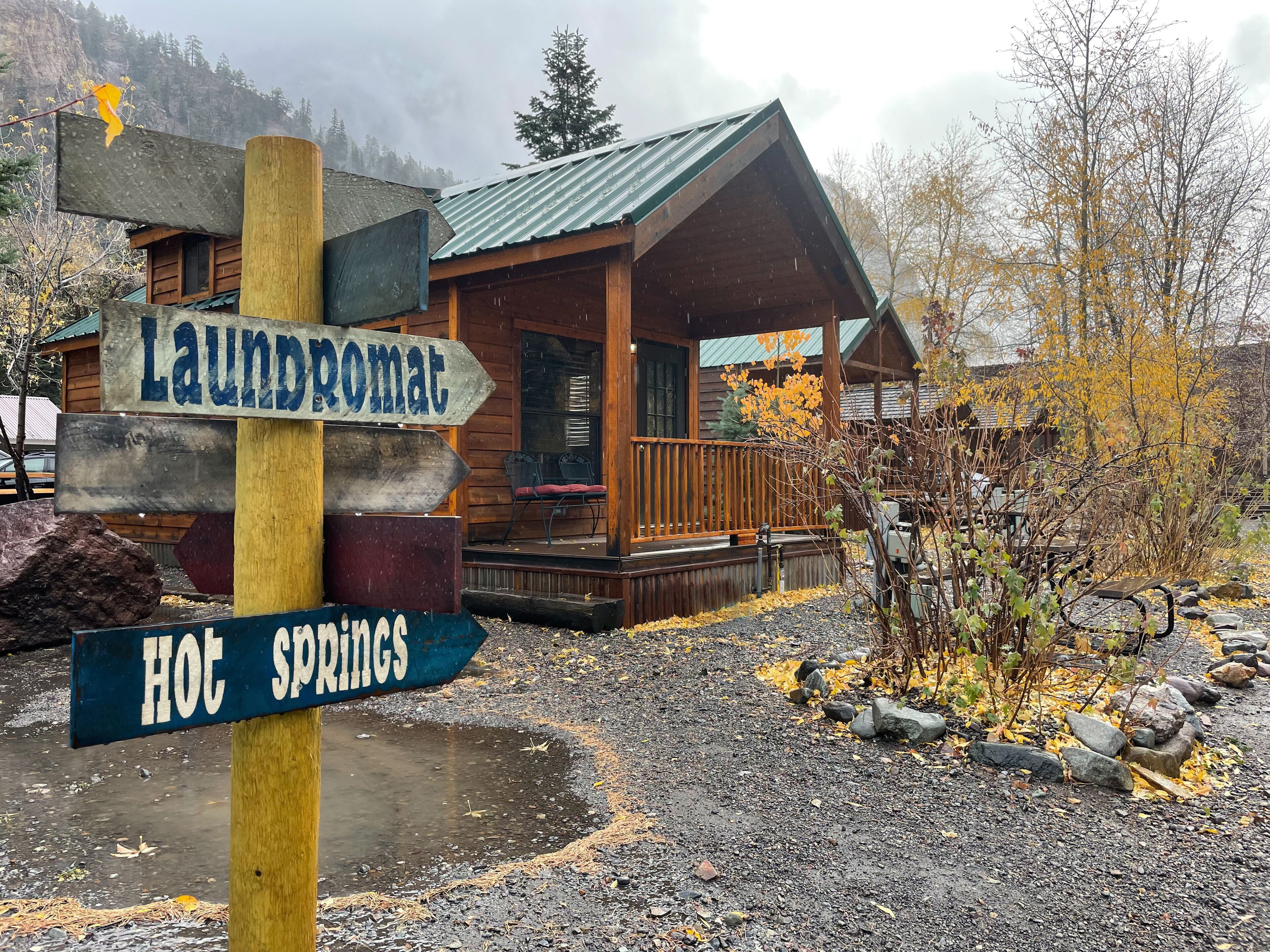 A wooden cabin with a porch, a sign indicating 'laundromat' and 'hot springs', and a mountainous backdrop.