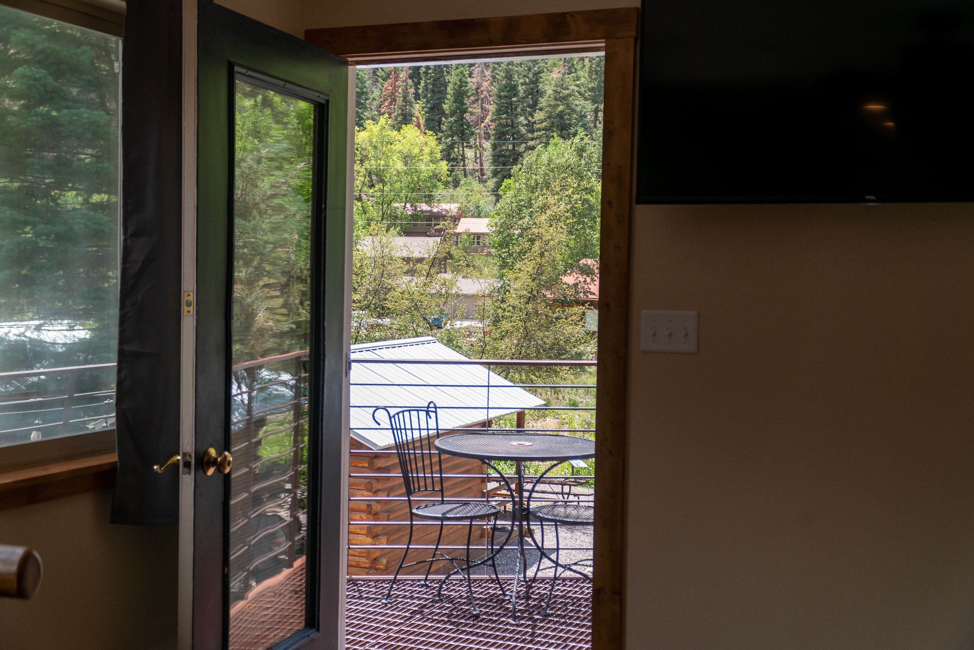 A room with a sliding glass door leading to an outdoor patio with a table and chairs, a TV mounted on the wall, and a view of a forested area.
