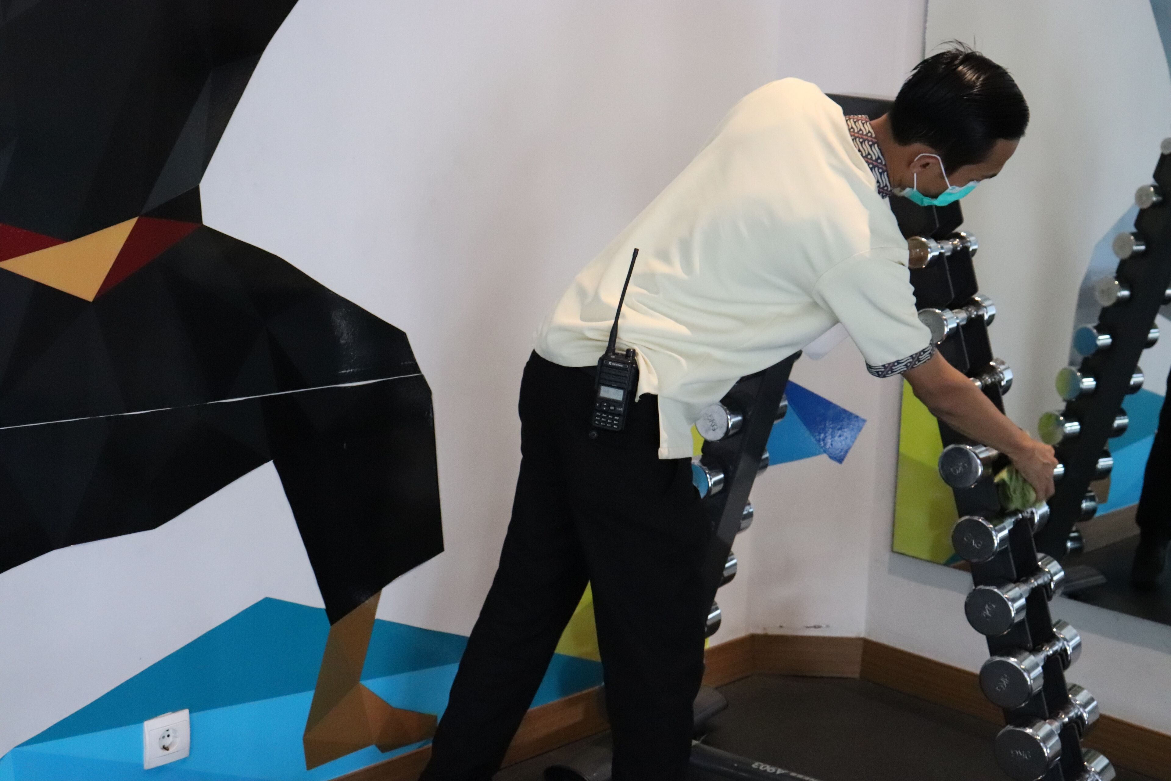 A person adjusting a row of dumbbells in a gym with geometric wall art and a mirror.