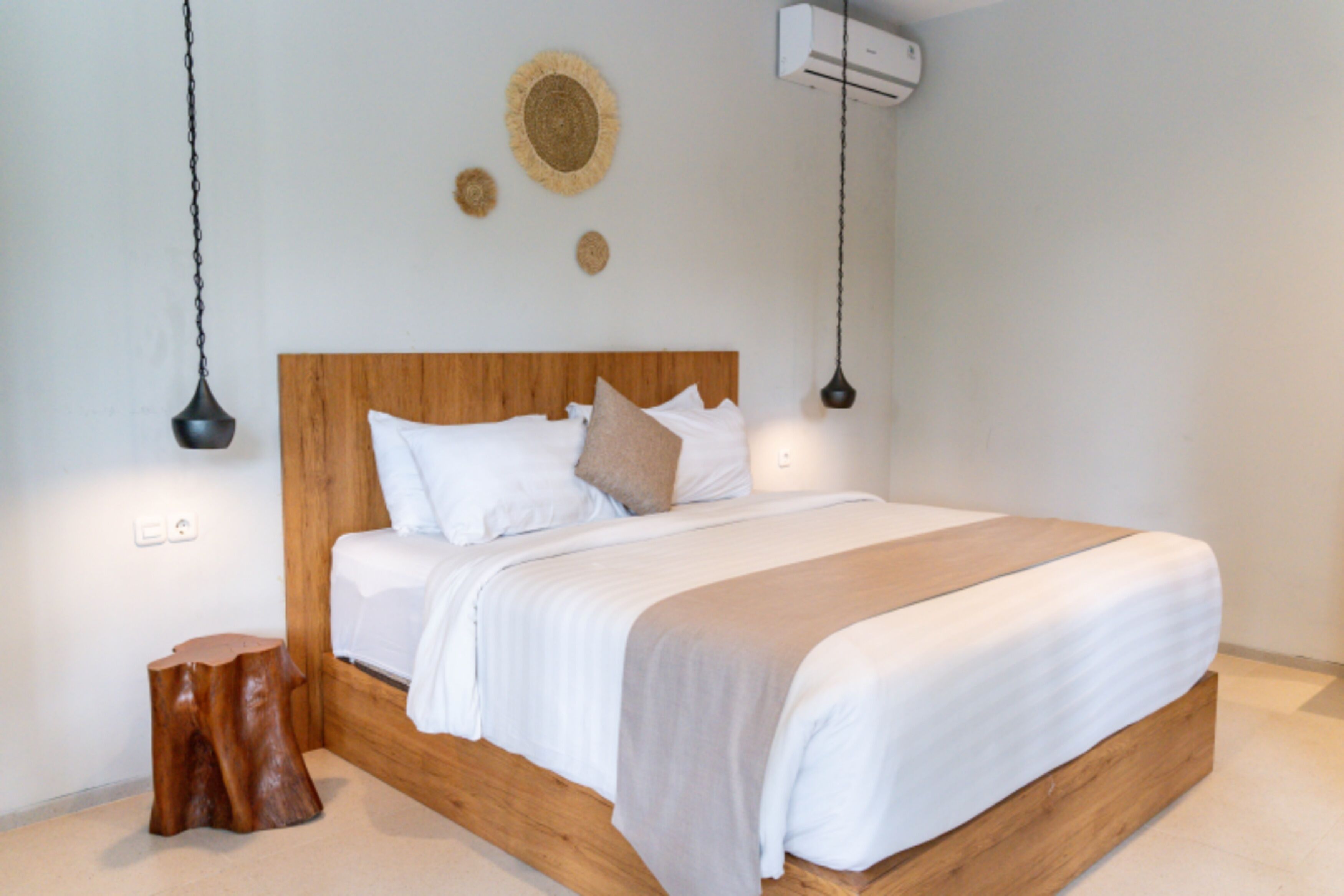 A bedroom with a wooden headboard, white bedding, a wooden stool, and a wall-mounted air conditioner.