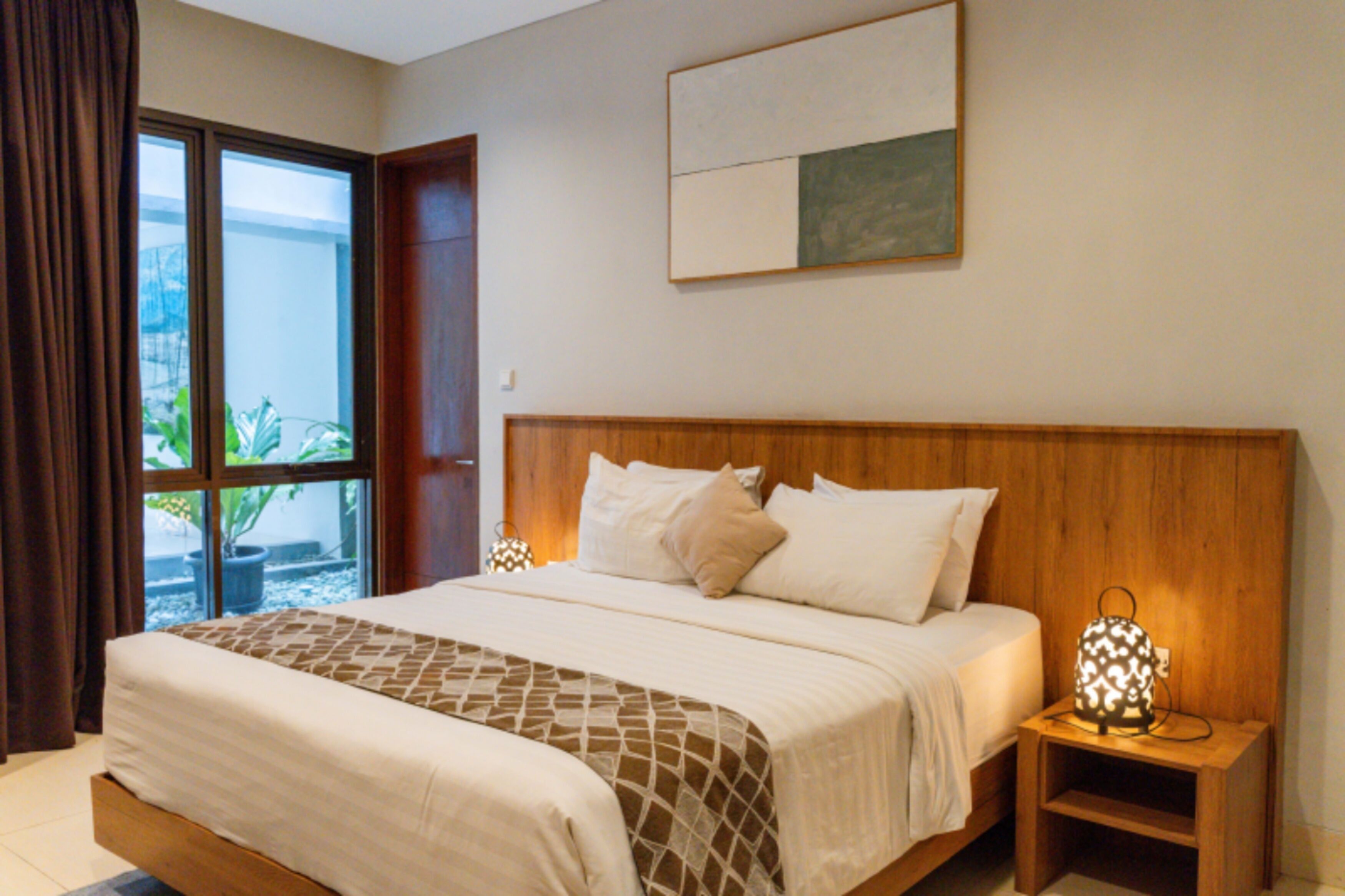 Een slaapkamer met een groot bed, een houten hoofdbord, een nachtkastje met een lamp en een raam met gordijnen.