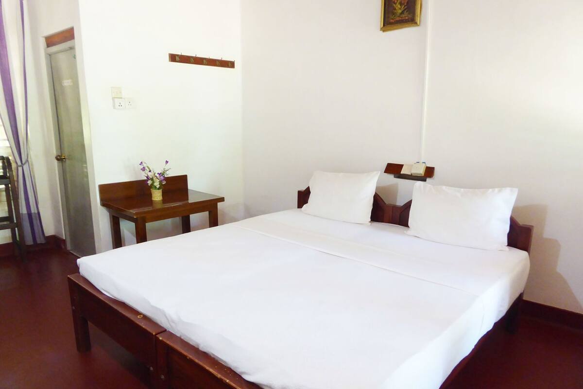 A neatly made bed with white linens, a wooden nightstand with a vase of flowers, and a small table with a chair.