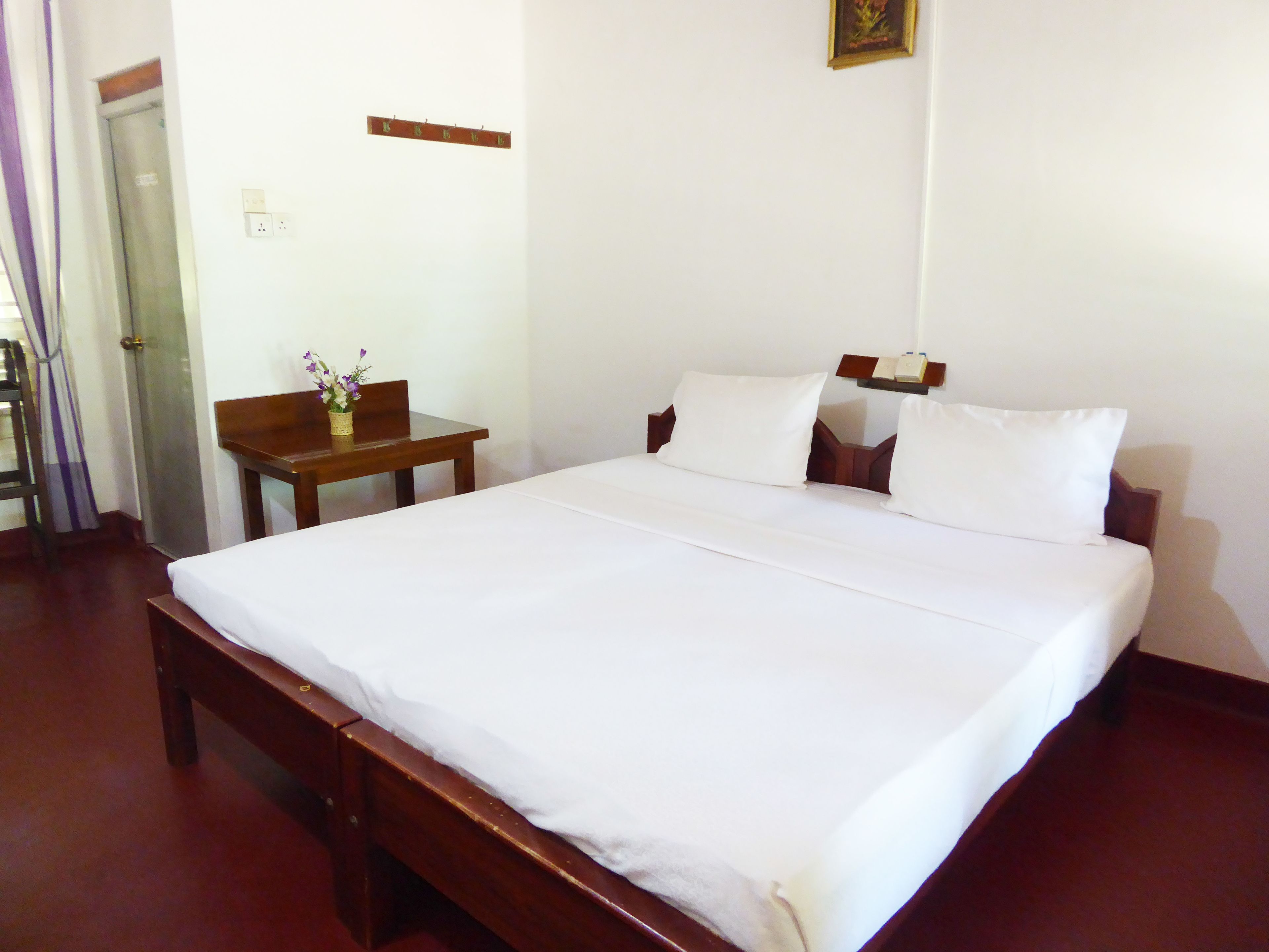 A neatly made bed with white linens, a wooden nightstand with a vase of flowers, and a wooden bench.