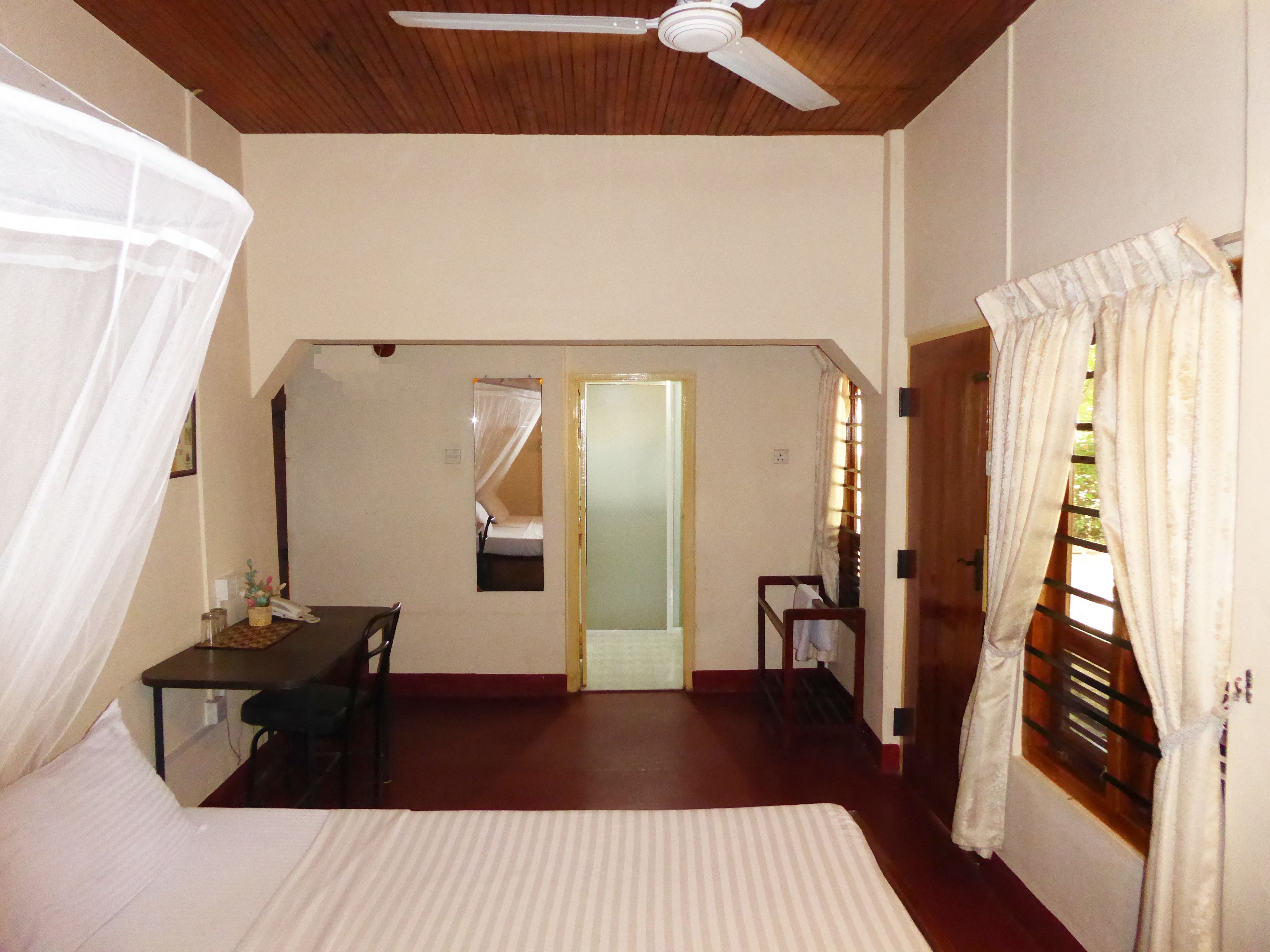 Een hotelkamer met een bed, een bureau, een stoel, een plafondventilator en een deur die naar een andere kamer leidt.