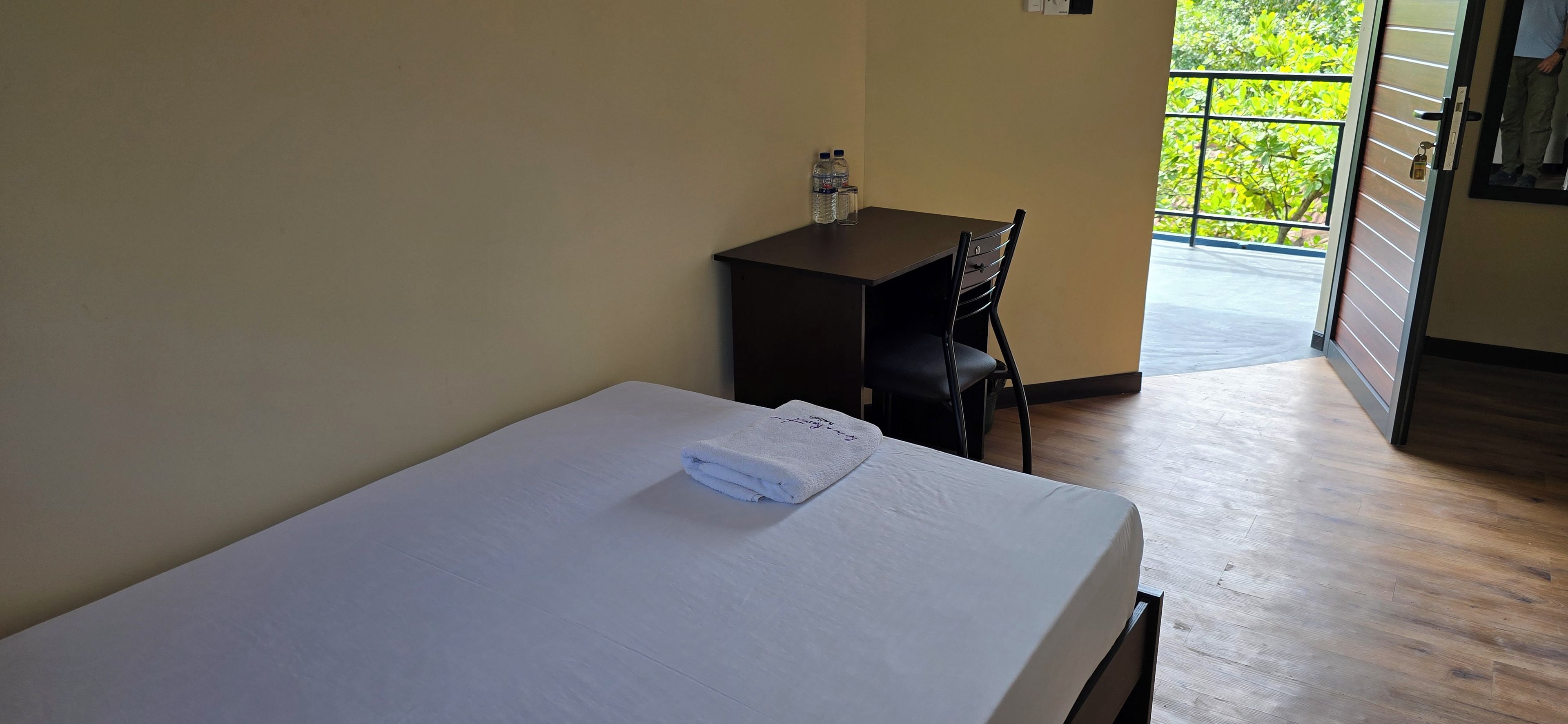 A hotel room with a bed, a desk, a chair, and a view of greenery outside.