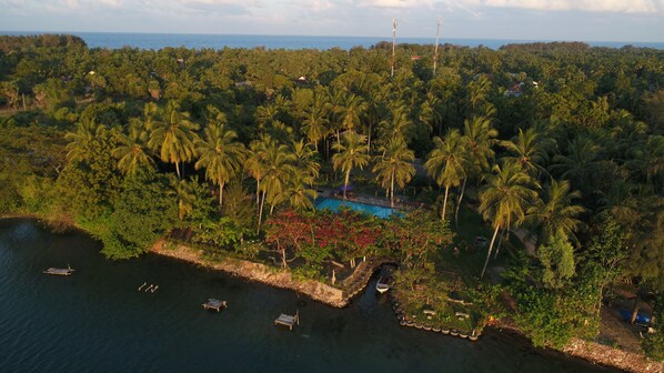 An aerial view of a tropical resort with a swimming pool surrounded by palm trees, boats on the water, and a distant view of the sea.