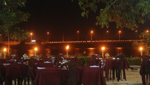 An outdoor dining area with tables set for guests, surrounded by trees and a bridge in the background. - Riviera Resort (Manmunai North)