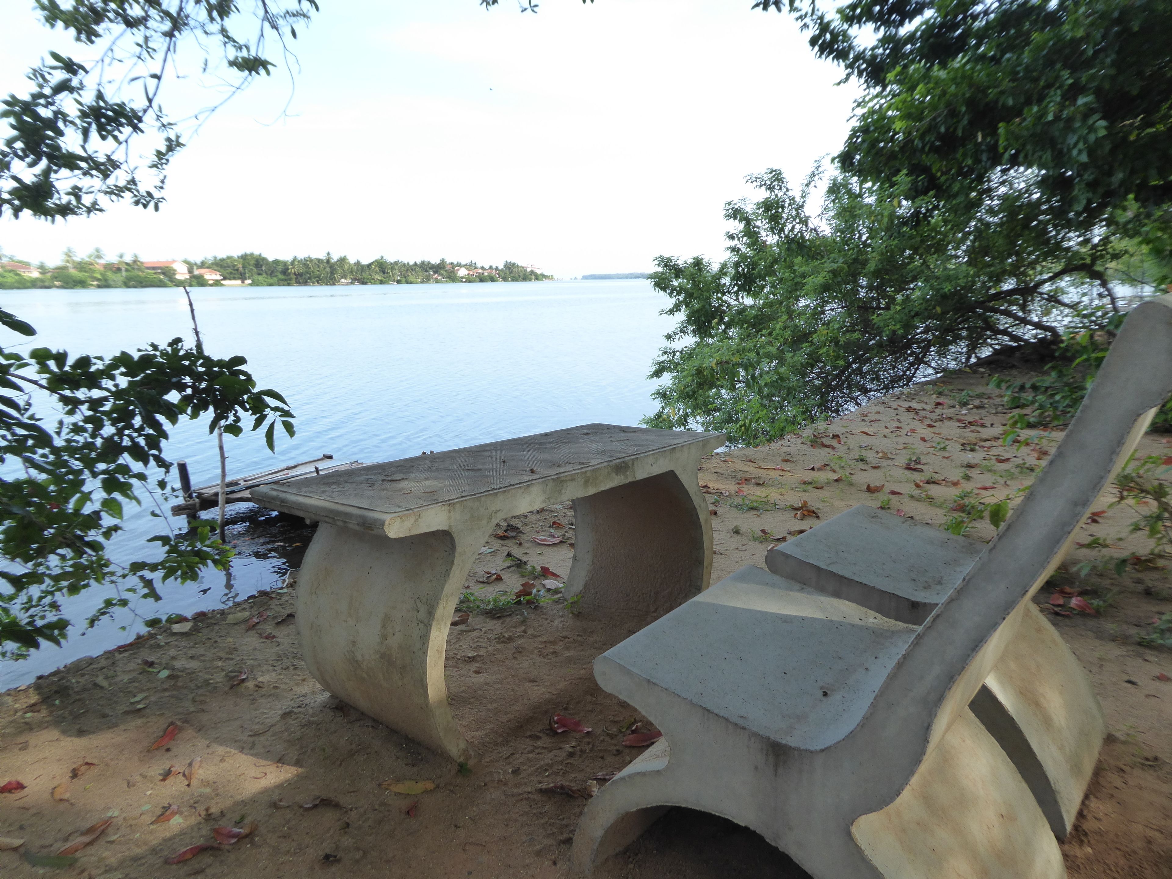 Betoninen penkki järven rannalla, jossa on hiekkaranta ja taustalla näkyy puita.