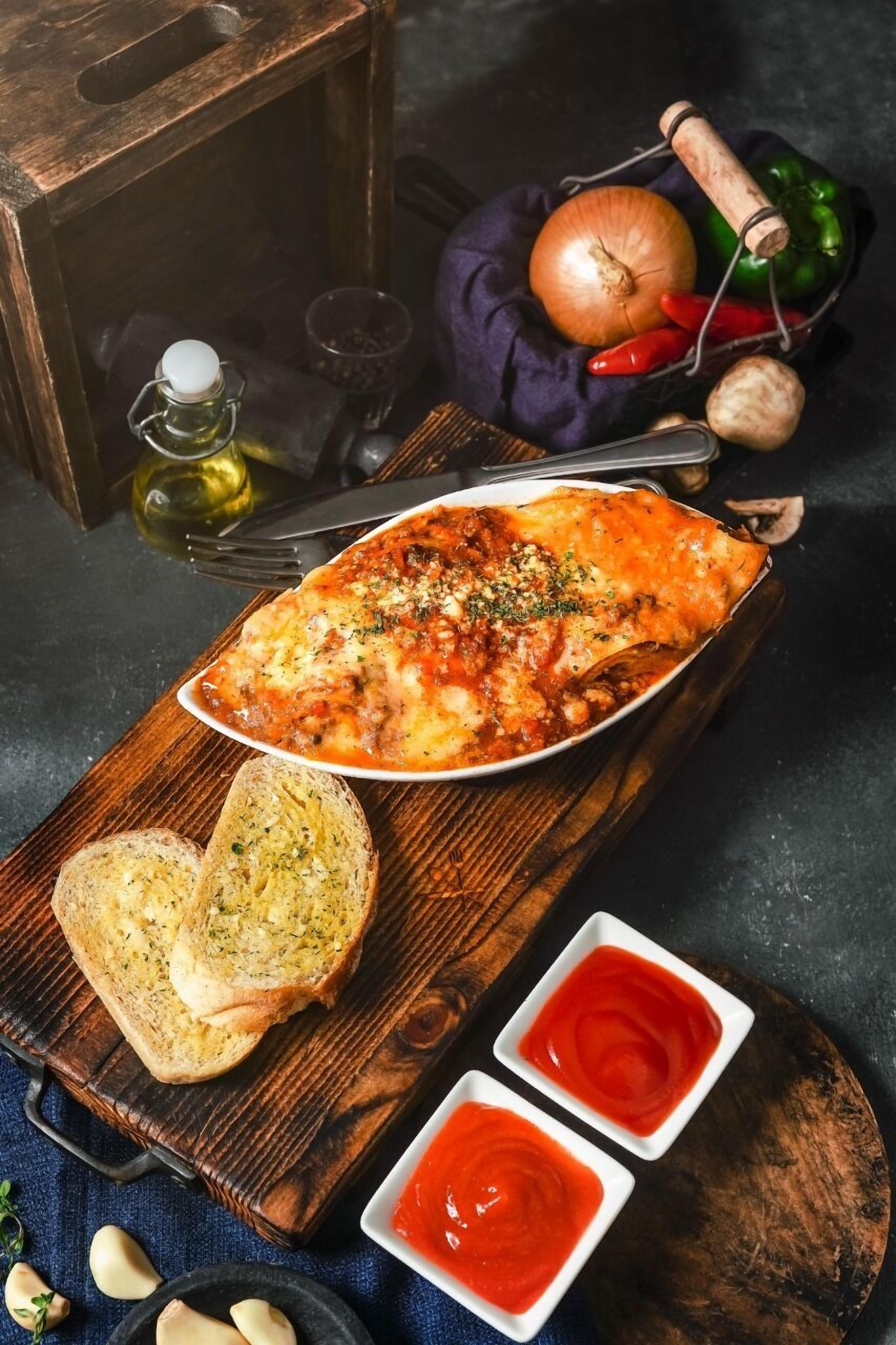 A wooden serving board with a dish of lasagna, two slices of garlic bread, and two small bowls of red sauce.
