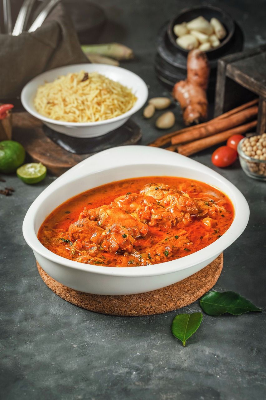 A bowl of curry with a side of rice, a lime, and some spices on a table.