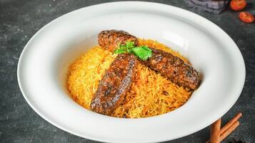 Ein weißer Teller mit einer Reisportion und zwei gegrillten Würstchen, verziert mit einem grünen Blatt, auf einer dunklen Oberfläche.