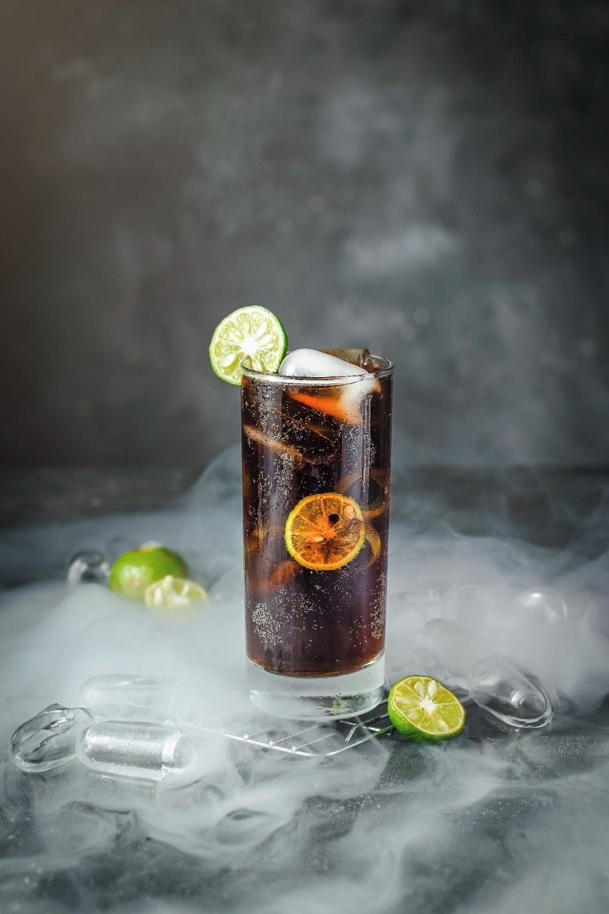 A tall glass with a dark beverage, ice, and lime slices, surrounded by ice cubes and a misty background.